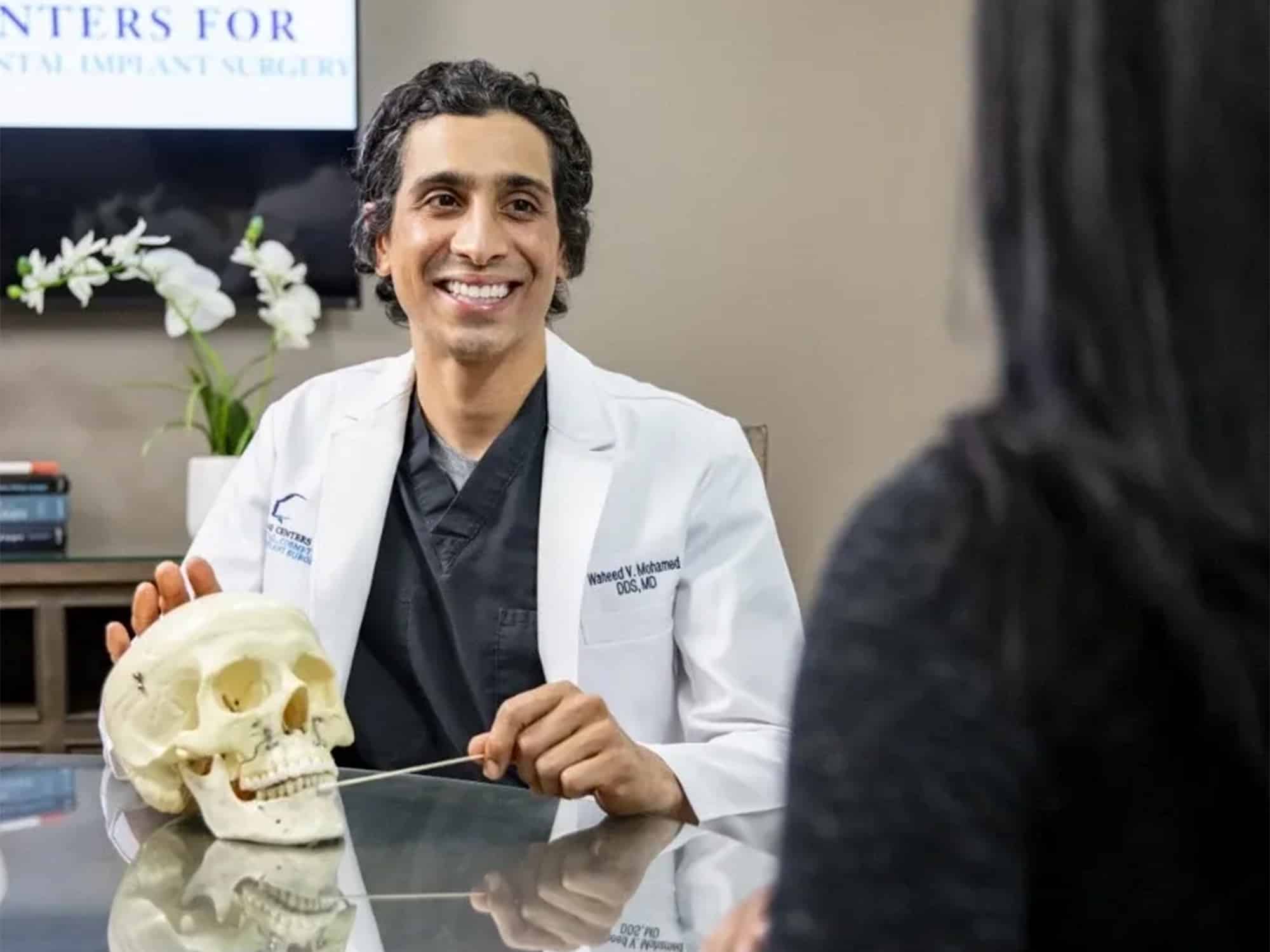 Two people in a dental office, discussing an anatomical skull model. A person in a white coat explains something with a pointer.