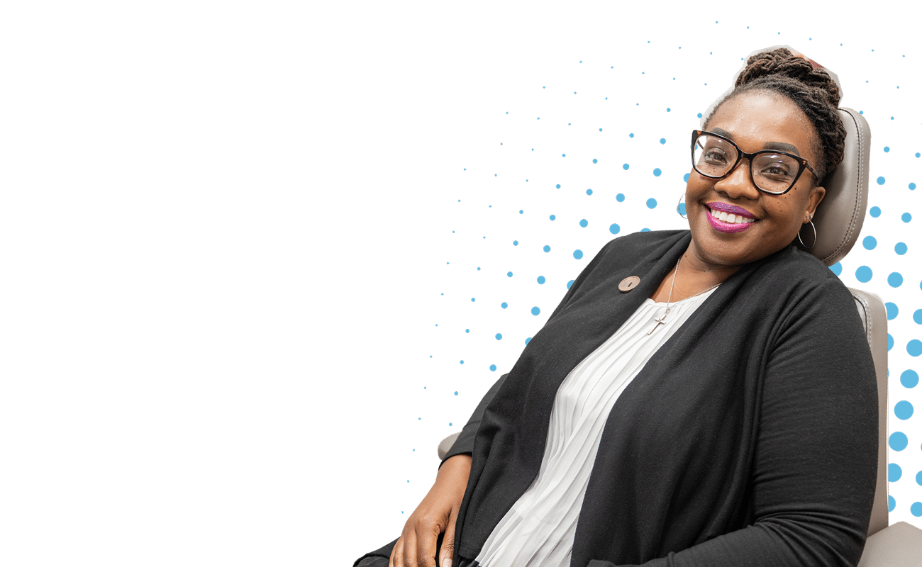 A person seated, smiling, wearing glasses and black attire, against a dotted black background, conveying a professional and cheerful demeanor.