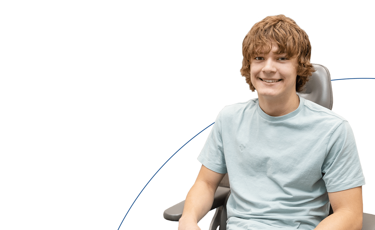 A person wearing a light blue shirt sits on a gray chair against a blue and white background, smiling at the camera.