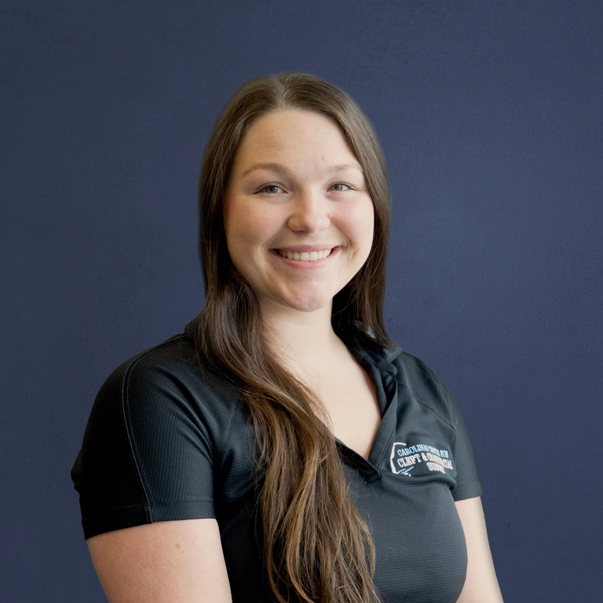 A person with long hair smiles in front of a plain, dark blue background, wearing a black shirt with text partially visible.