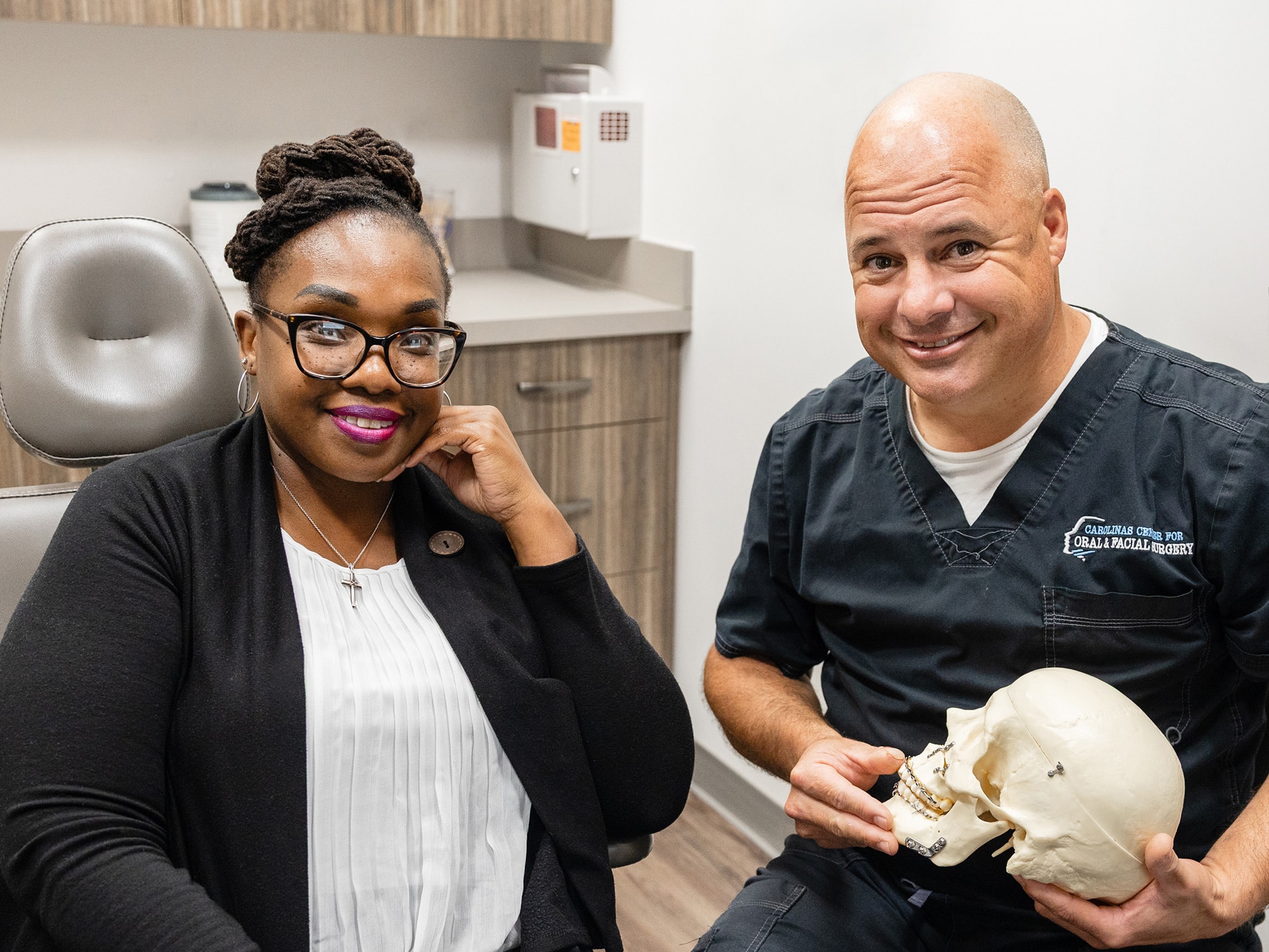 Two people in a medical office; one holds a skull model. They are smiling, seated by a dental chair and cabinets.