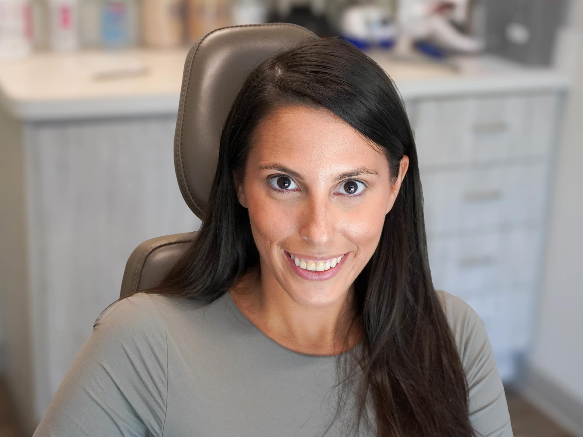 A person with long dark hair smiles while sitting on a chair in a modern office setting with cabinets in the background.