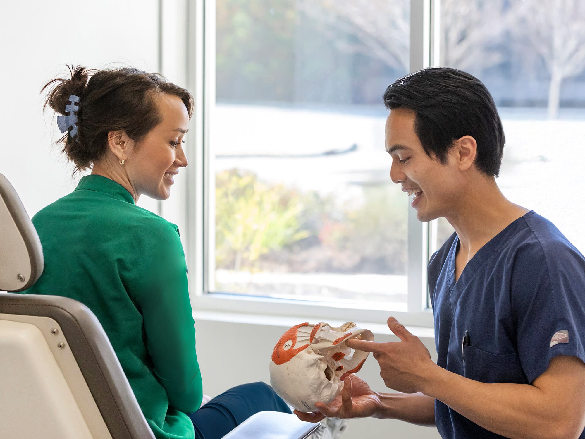 Two people are in a medical setting; one in scrubs explains a skull model. Bright window light enhances focus on conversation.