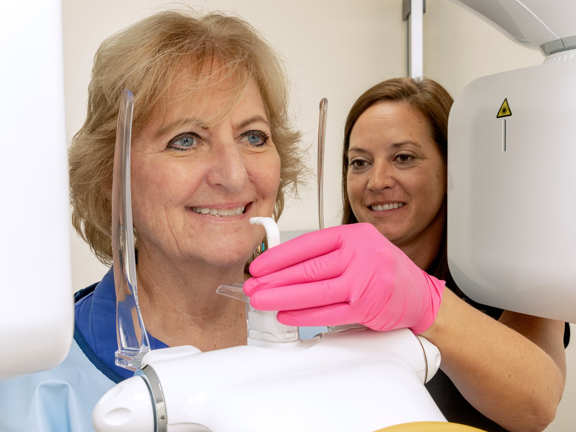 A person assists another in using dental imaging equipment. The person in gloves adjusts the machine for a dental X-ray or scan.