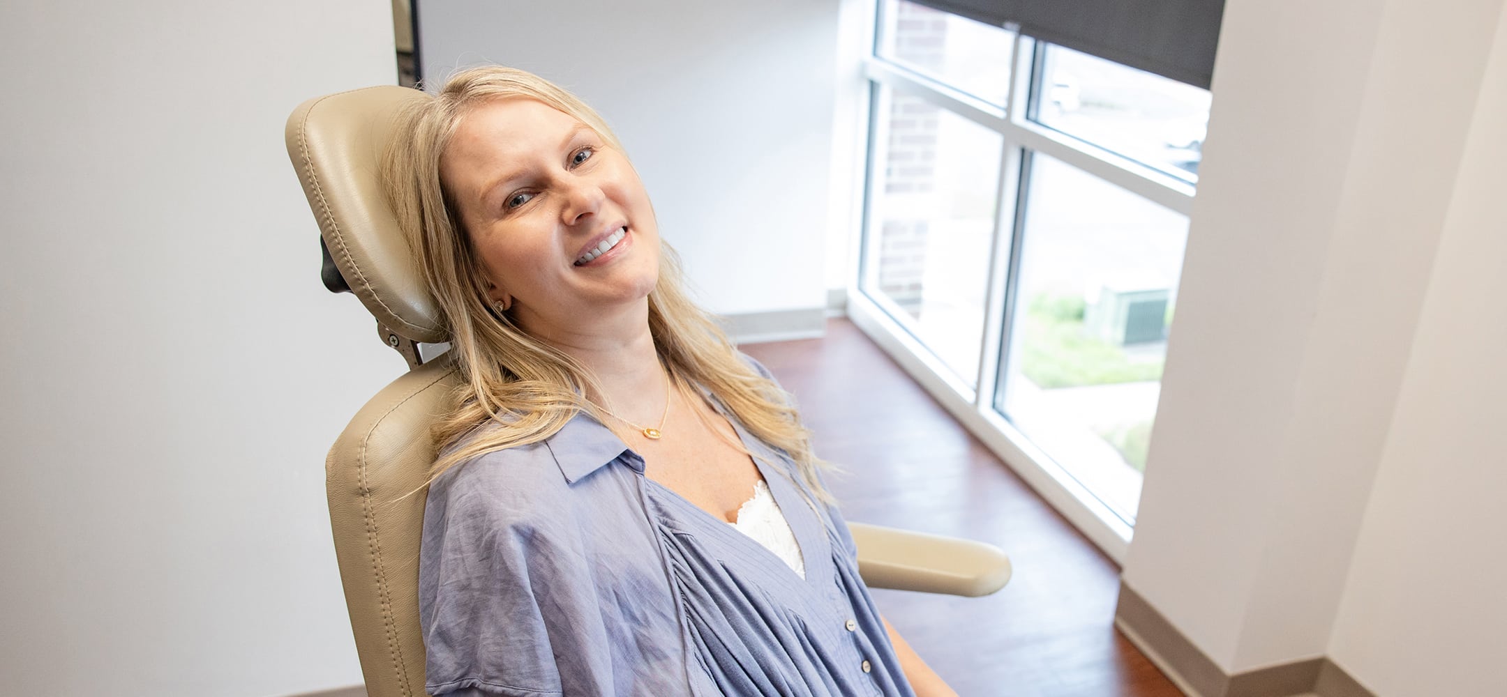 A person wearing a light blue shirt sits smiling in a medical chair by large windows, in a modern, bright room.