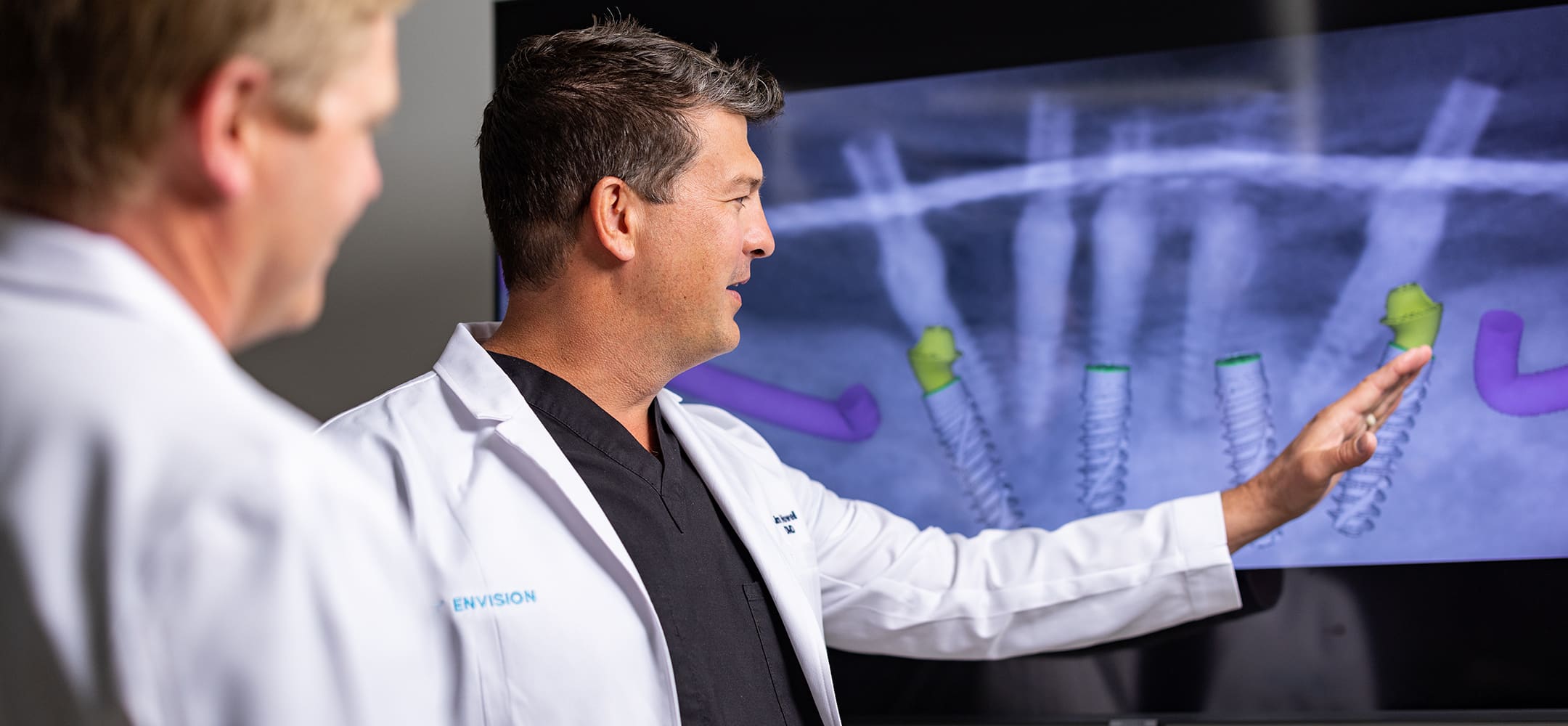 Two persons in lab coats review a dental X-ray on a screen, discussing the highlighted areas.