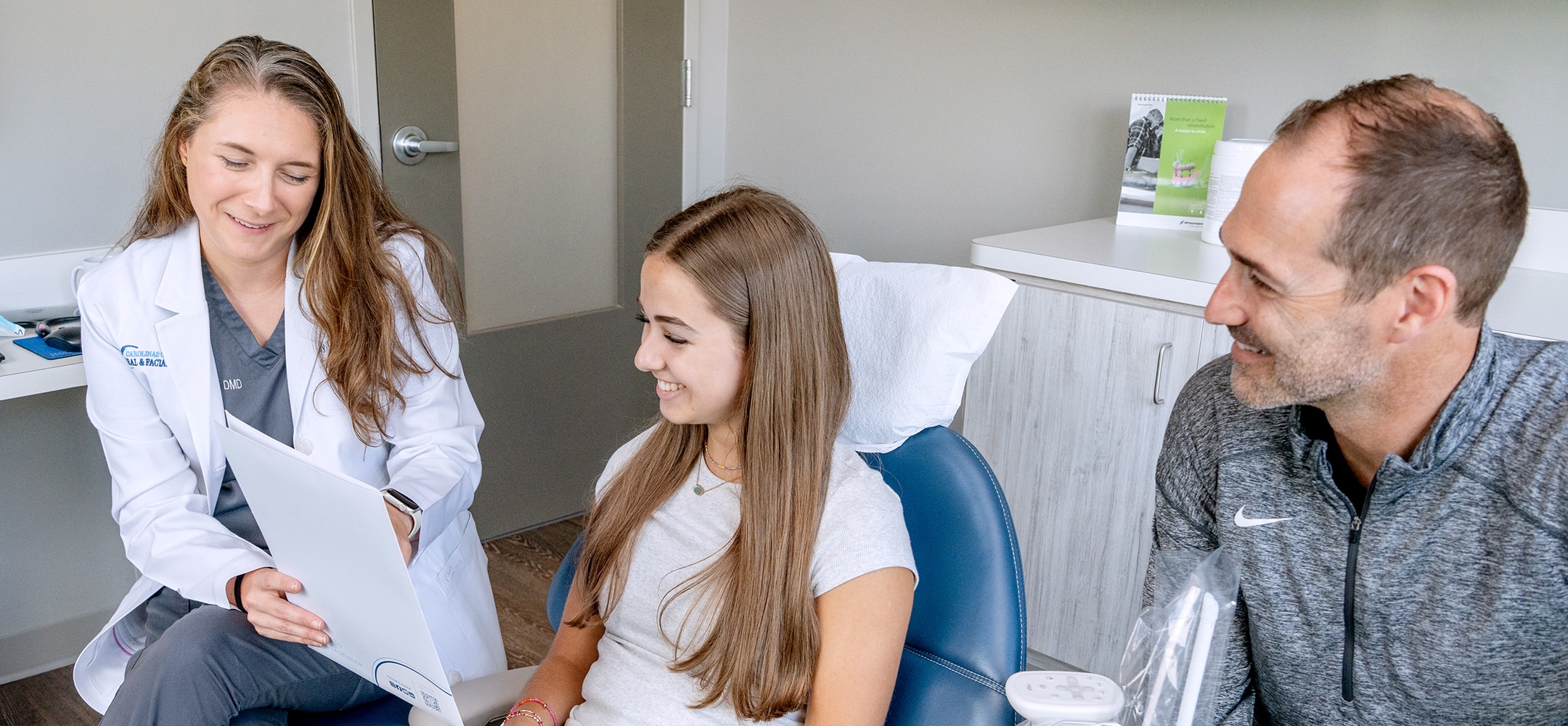 A person in a white coat discusses plans with a patient and another person in a modern clinic setting, all smiling.