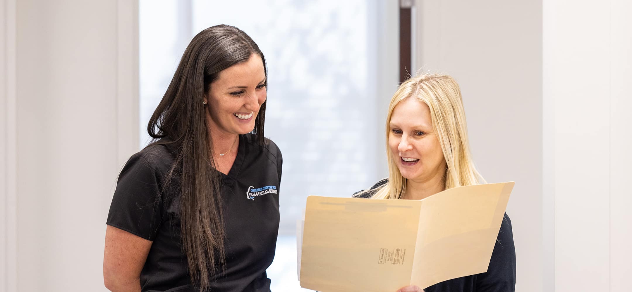Two people in a bright room, smiling and discussing a document. One holds a folder while both appear engaged and professional.