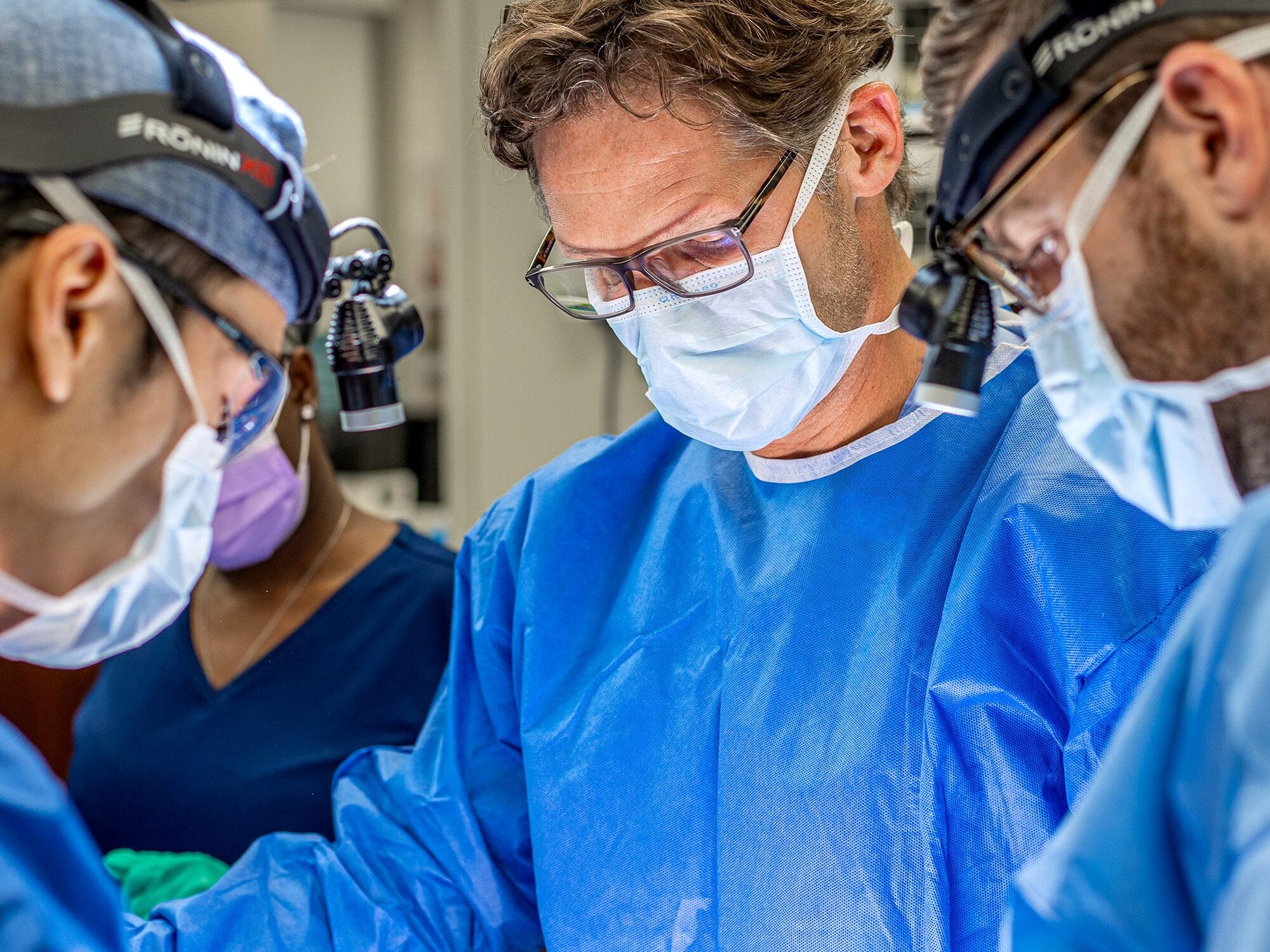 Four medical professionals in blue surgical attire focus intently during a procedure, wearing masks and headlamps in a well-lit operating room.