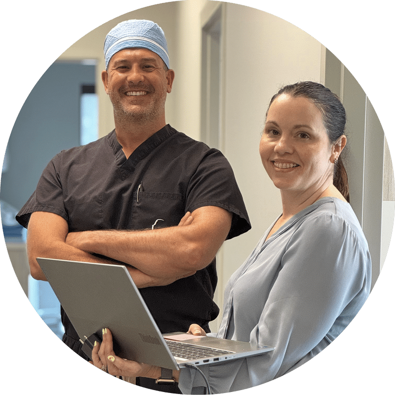 Two smiling people in a hallway, one in scrubs with a cap, the other holding a laptop, standing side by side indoors.