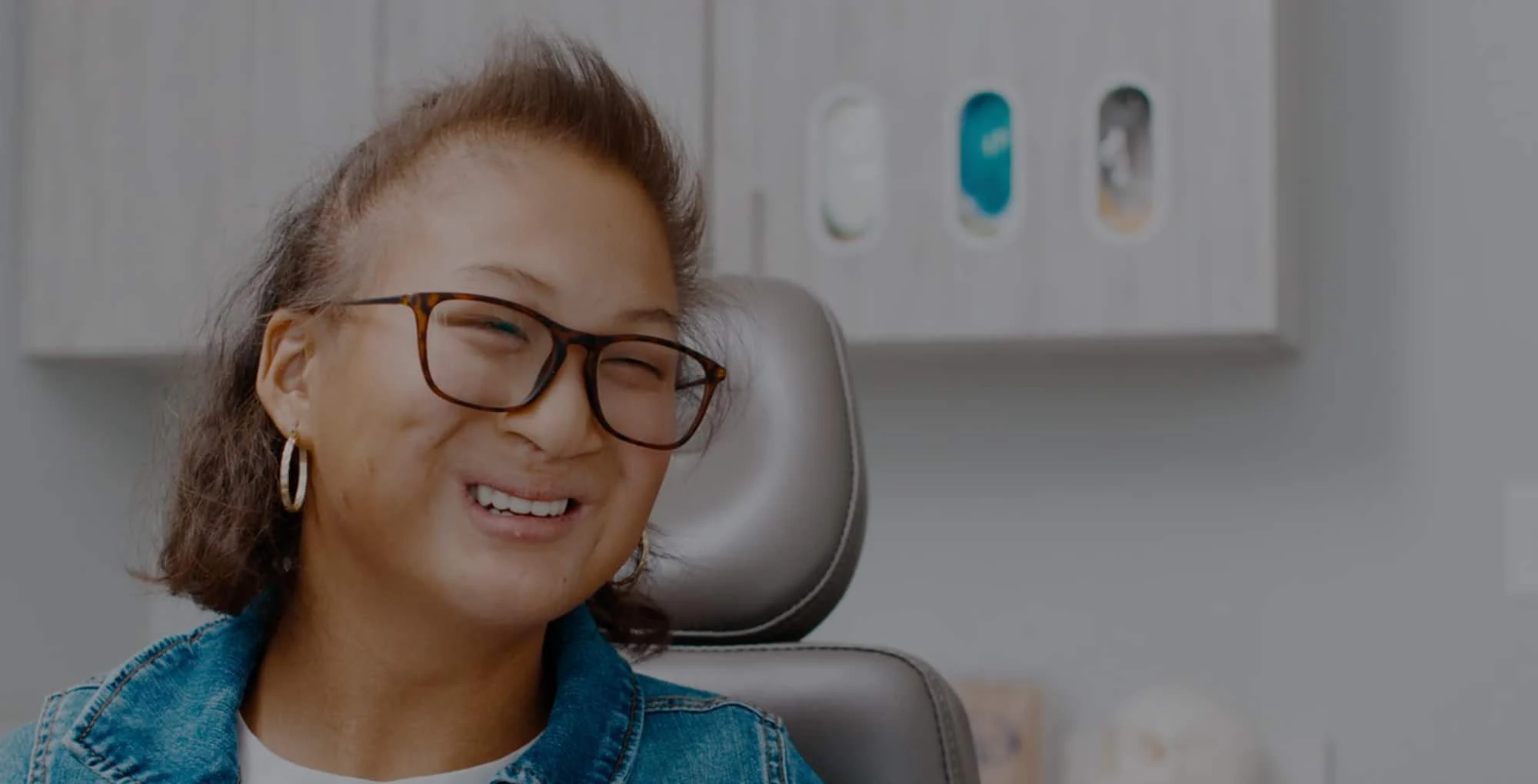 A smiling person wearing glasses and a denim jacket sits in an office with display shelves in the background.