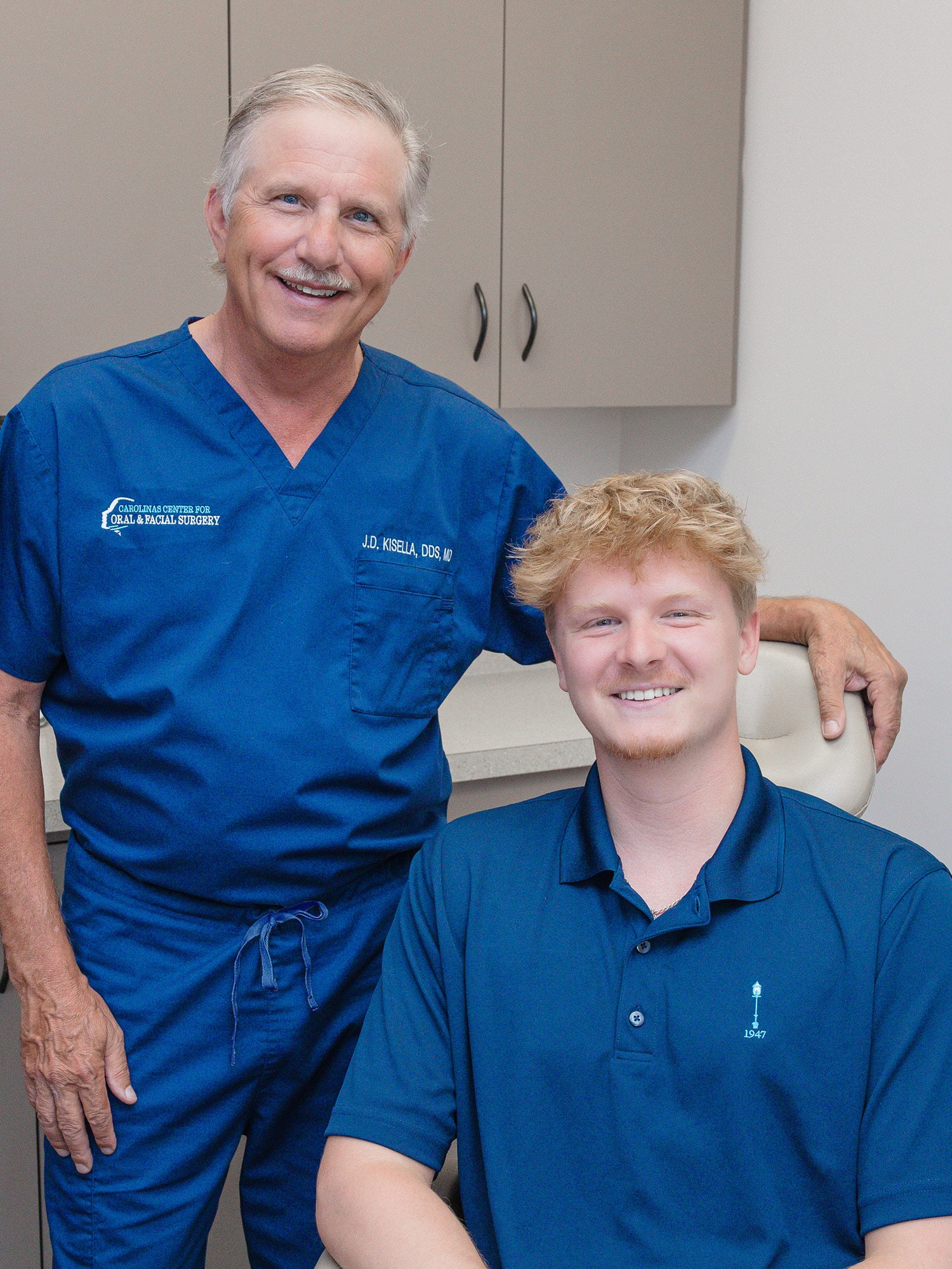 Two people in blue attire are smiling in an office setting. One is standing, the other seated. Cabinets are visible behind them.
