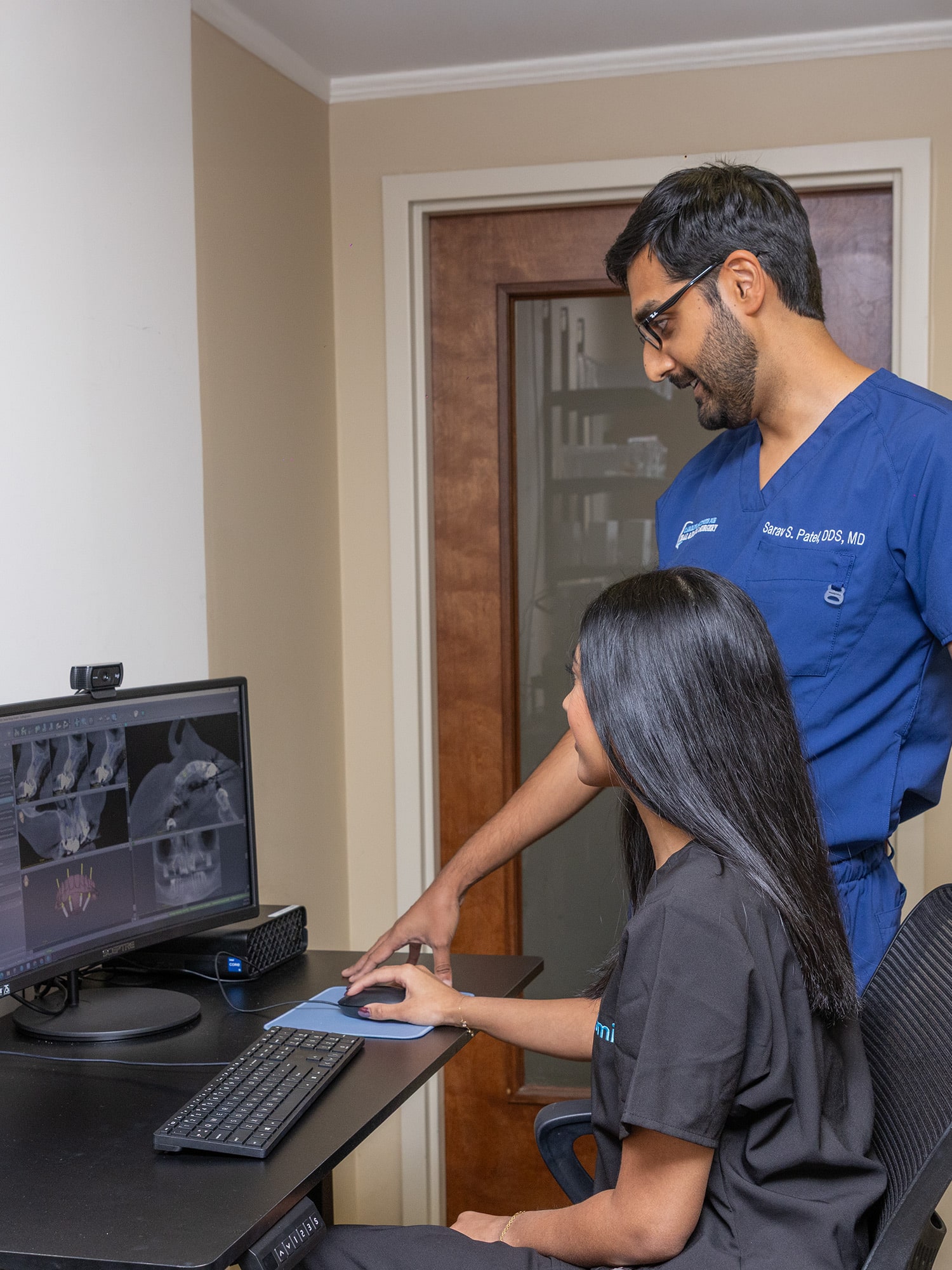 Two people in a medical setting examine dental X-rays on a computer monitor, both wearing scrubs, engaging in professional discussion.
