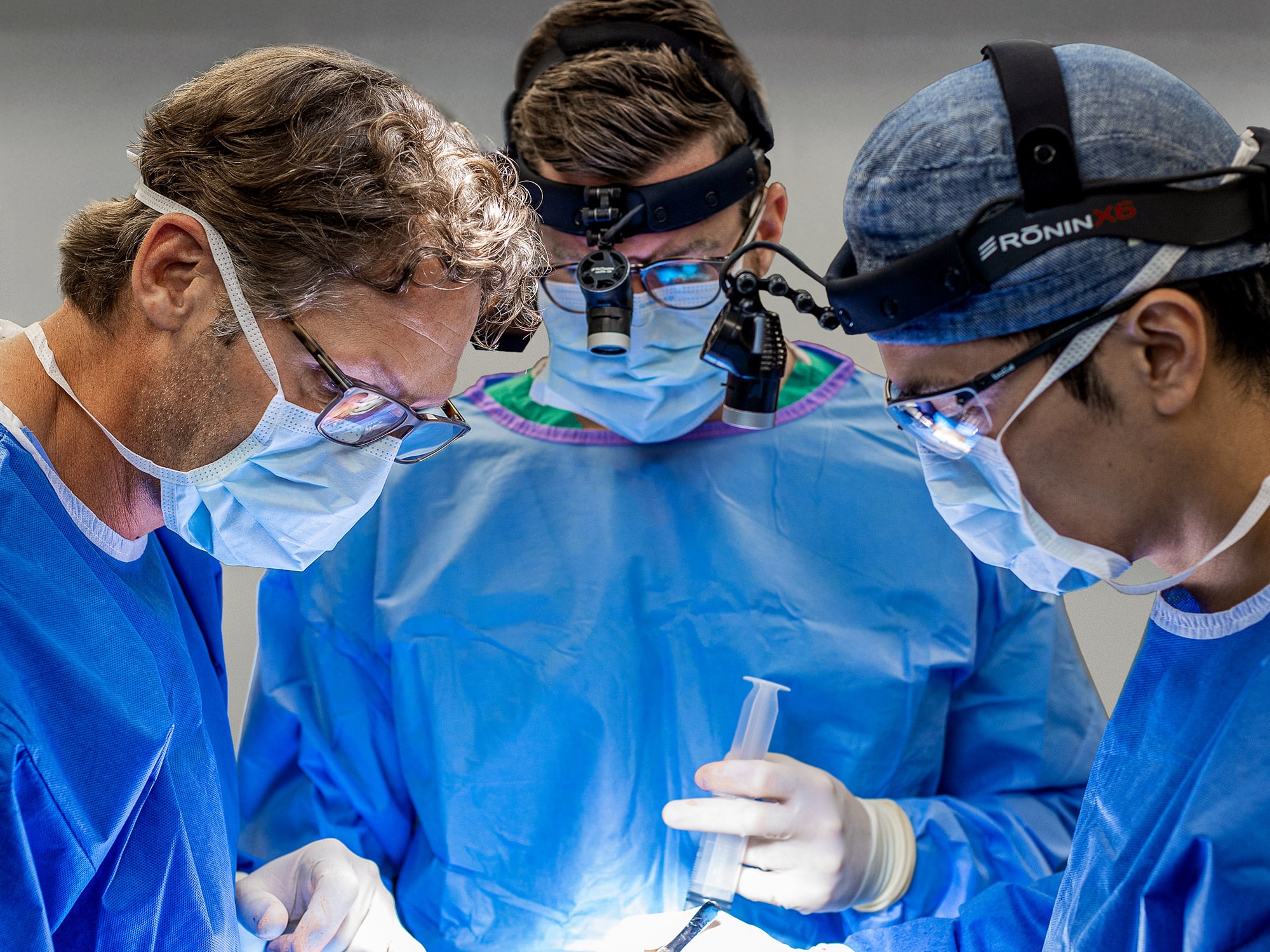Three medical professionals in surgical attire and masks focus intently on a procedure, using specialized tools under bright operating room lights.
