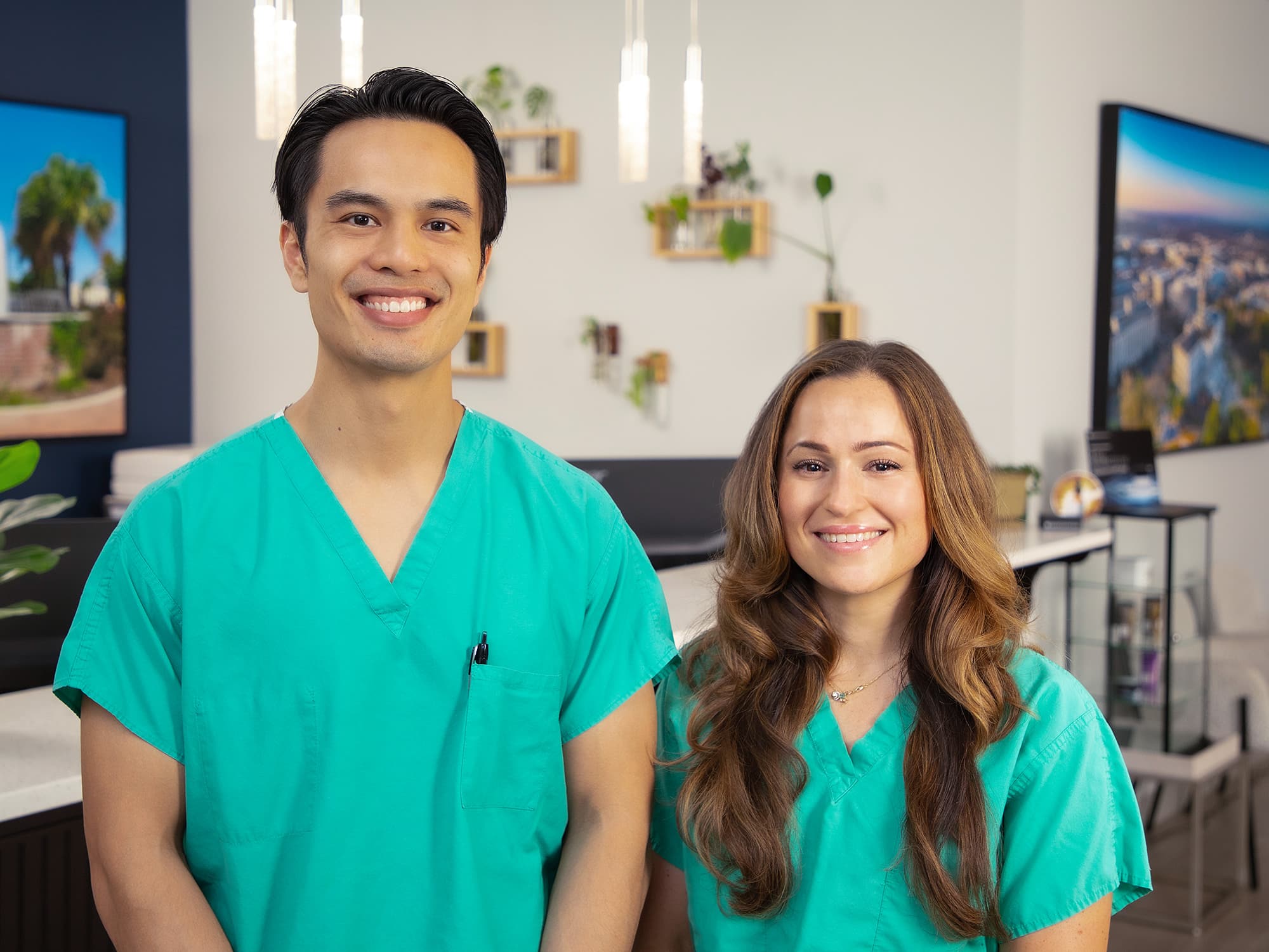 Two people in green medical scrubs smiling, standing indoors. Background features plants, cityscape artwork, and modern decor with hanging lights.