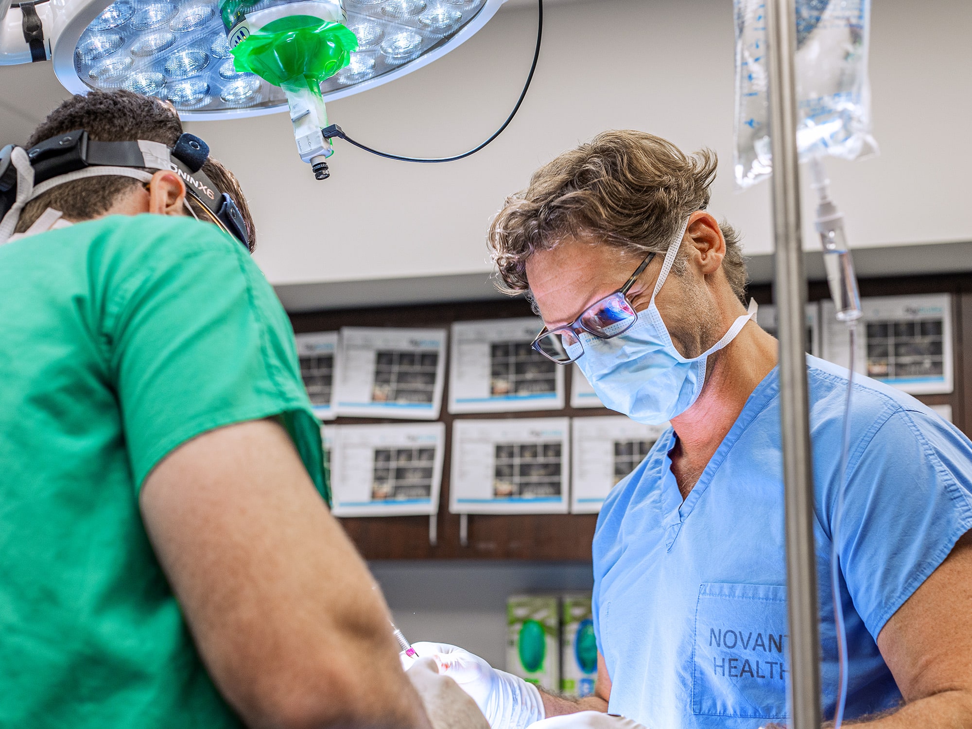 Two people in medical scrubs perform surgery under an operating room light. Surgical tools and IV equipment are visible in a clinical setting.