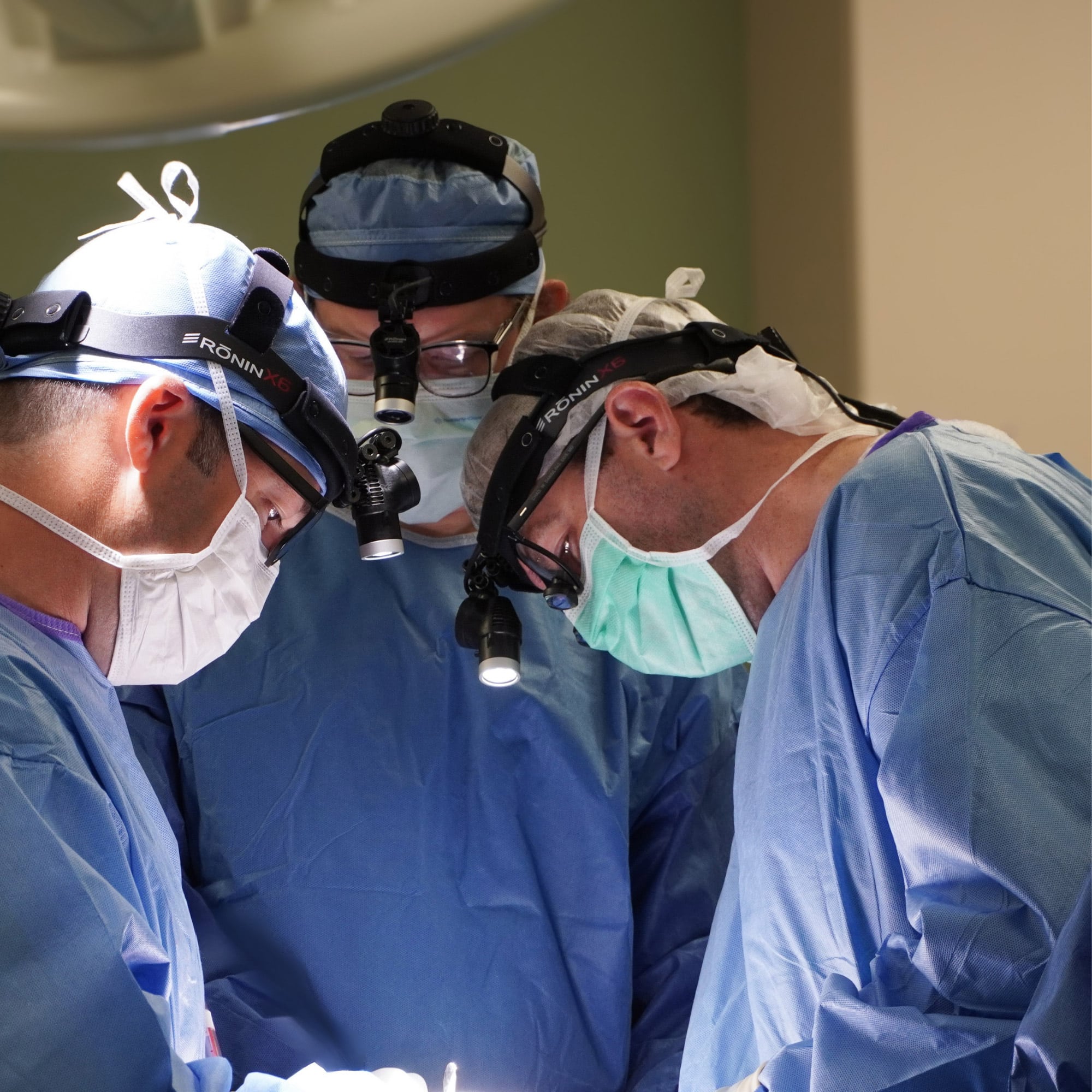 Three medical personnel, wearing blue surgical attire and headlamps, intensely focus on a procedure under bright operating room lights.