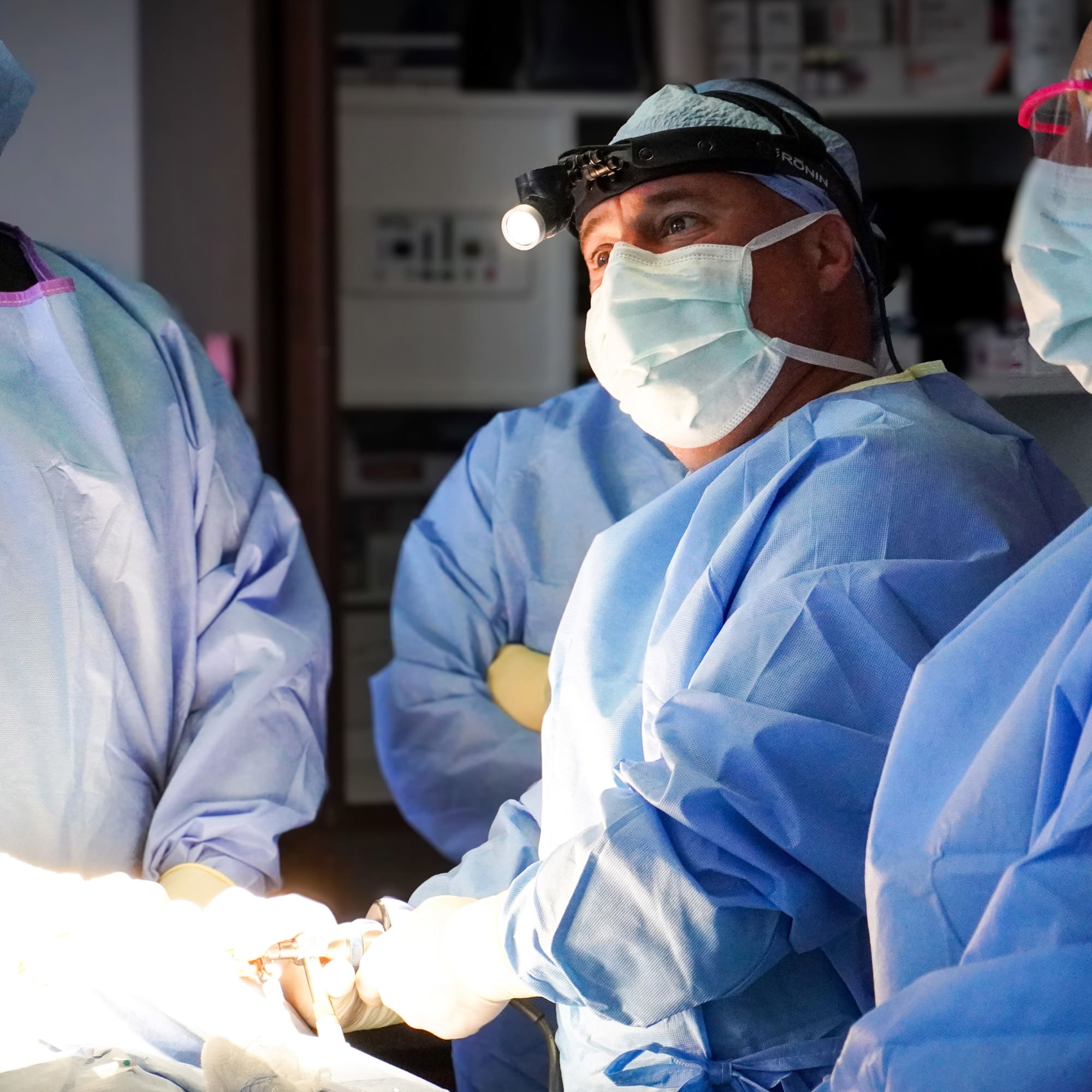 Medical team in blue scrubs performing surgery. One person wears a headlamp, focused on the procedure, in a clinical operating room setting.