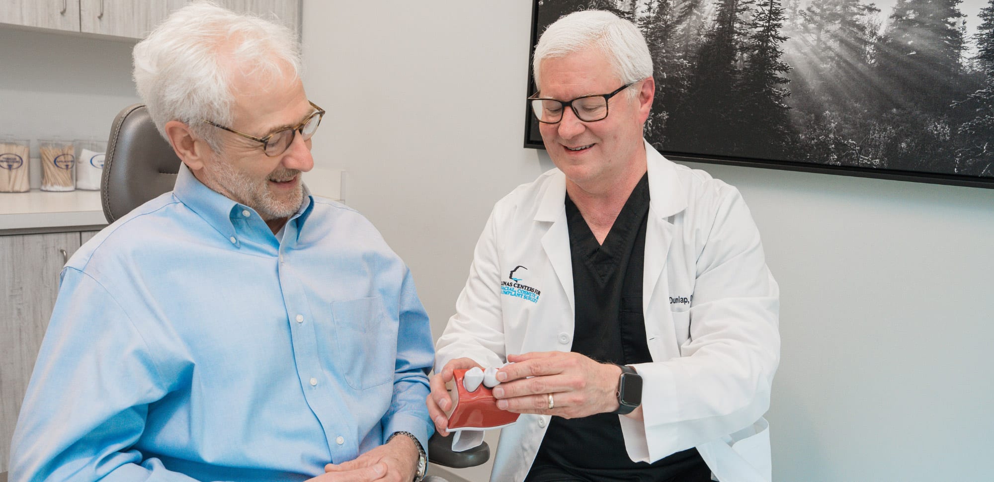 Two people in a medical office discuss dental health using a tooth model, with forest artwork in the background.