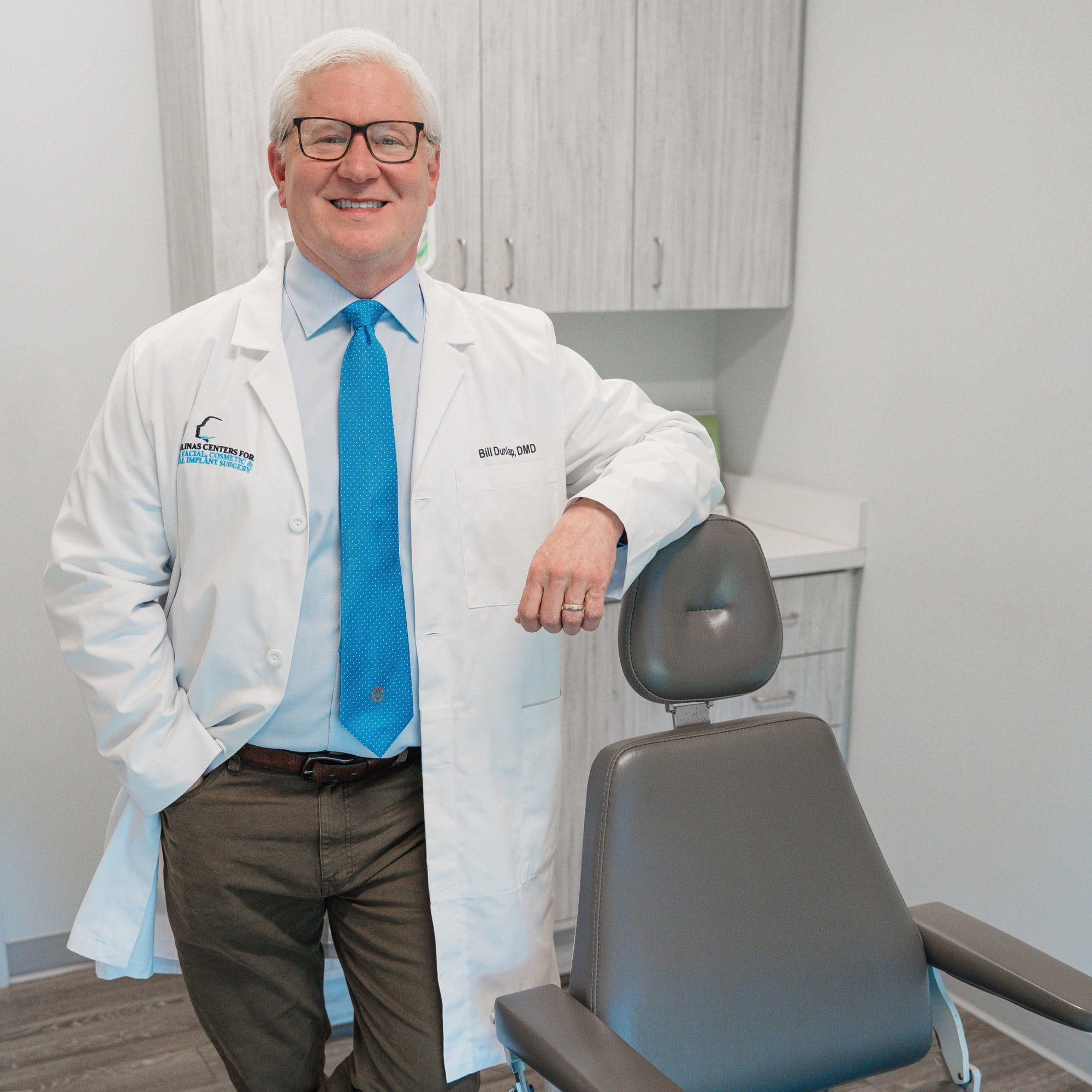 A person in a white coat stands beside a dental chair in a modern clinic room, smiling and wearing glasses and a blue tie.