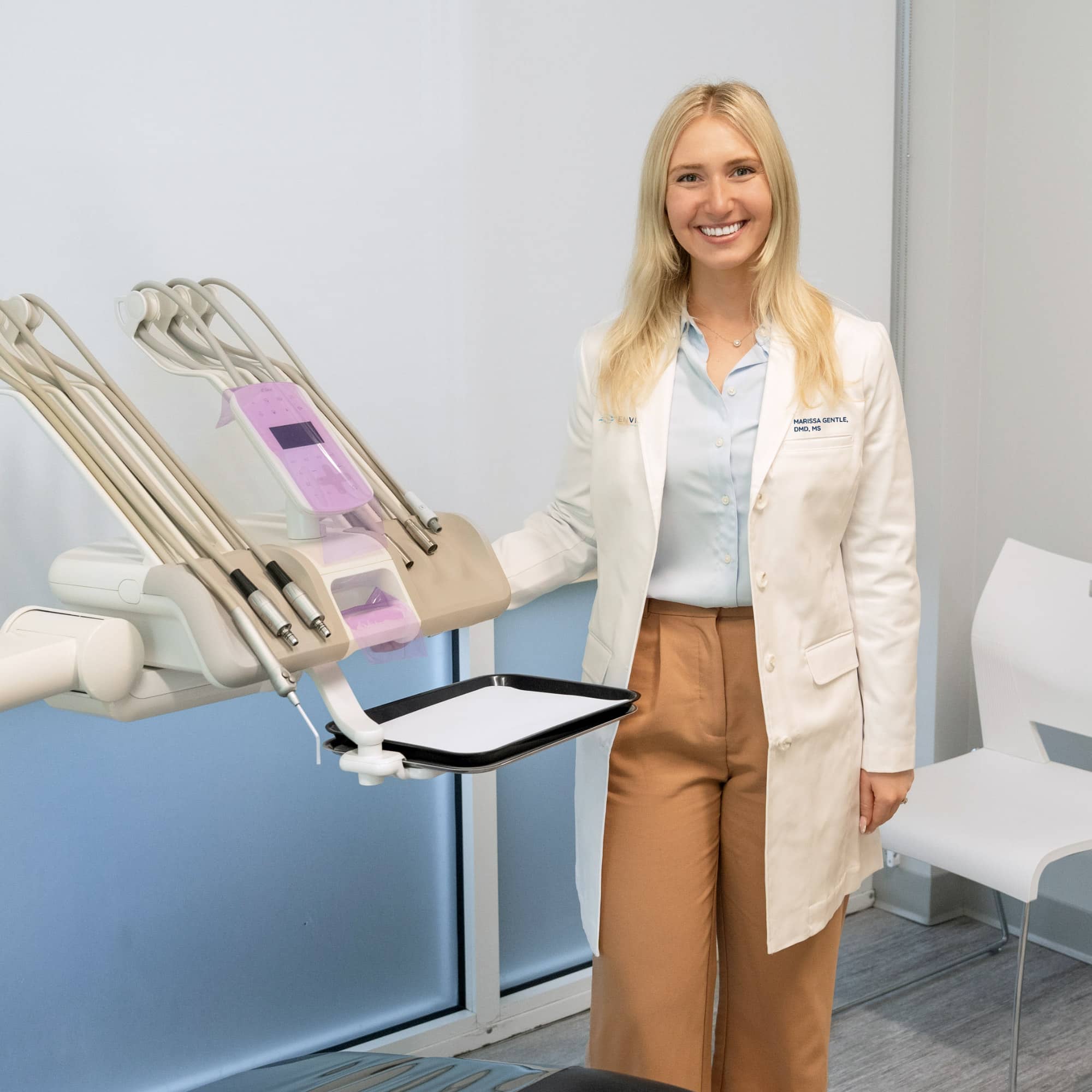 A person wearing a white coat stands in a dental office, smiling next to dental equipment, with a modern, clean design interior.