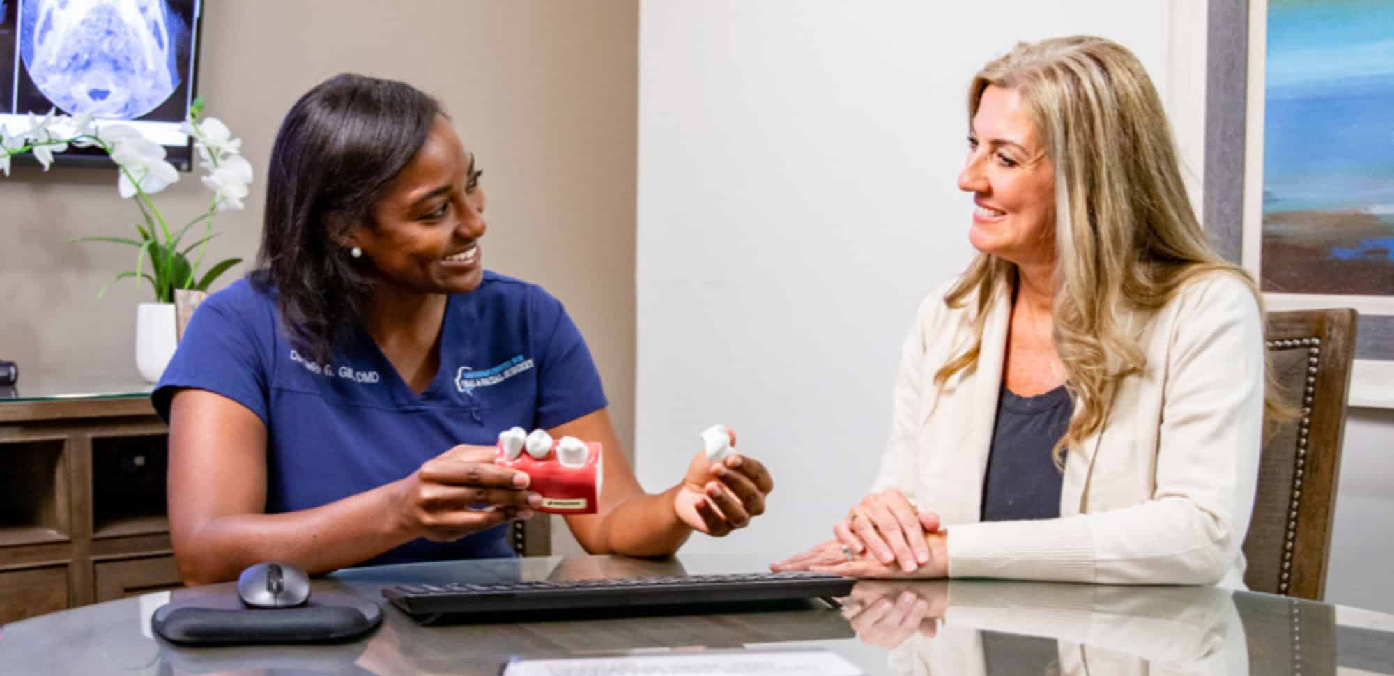A person in scrubs shows dental models to another person sitting at a desk, discussing dental care.