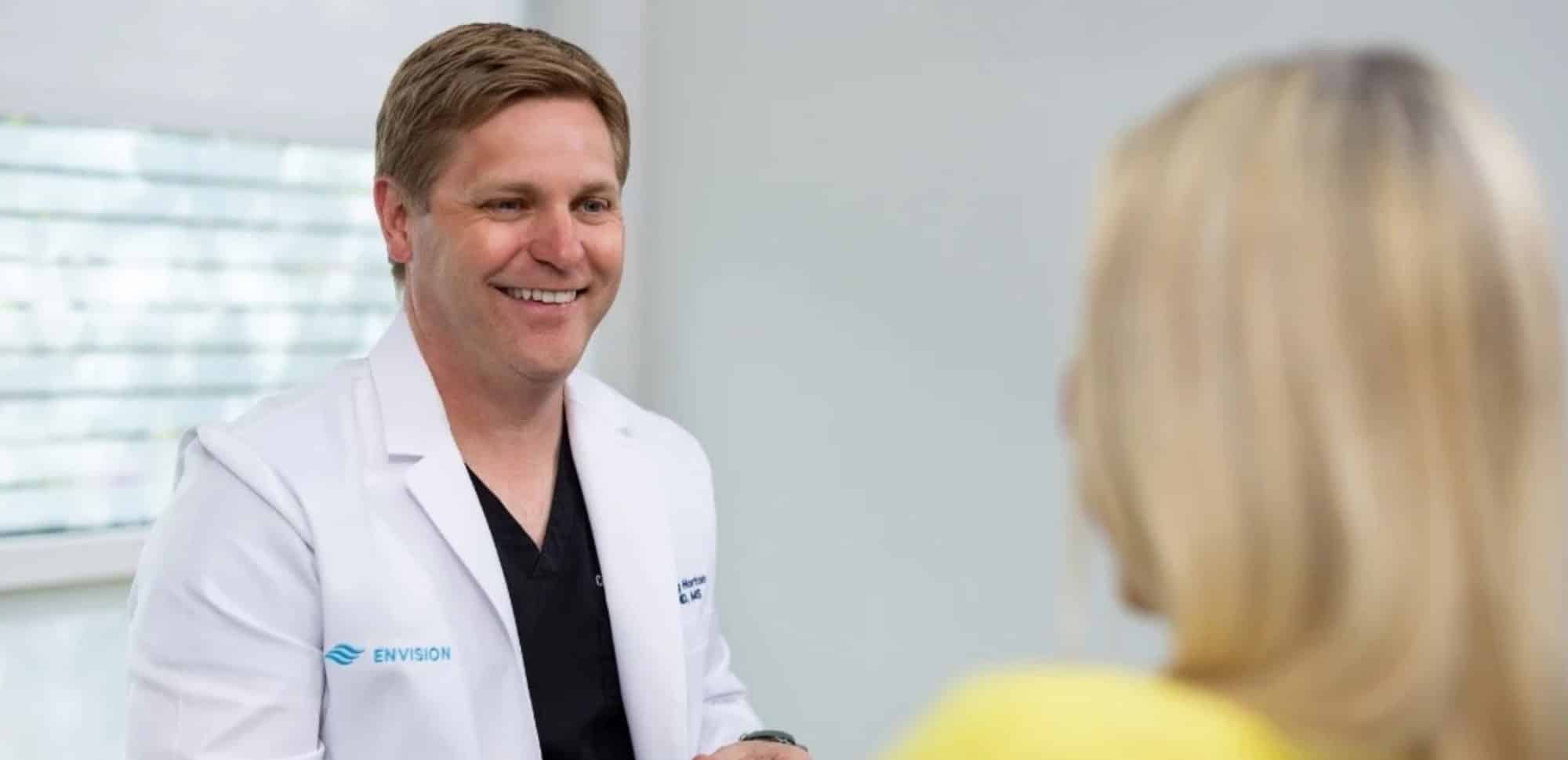 A person in a white coat smiles at another in a bright room. No landmarks or historical buildings are visible.