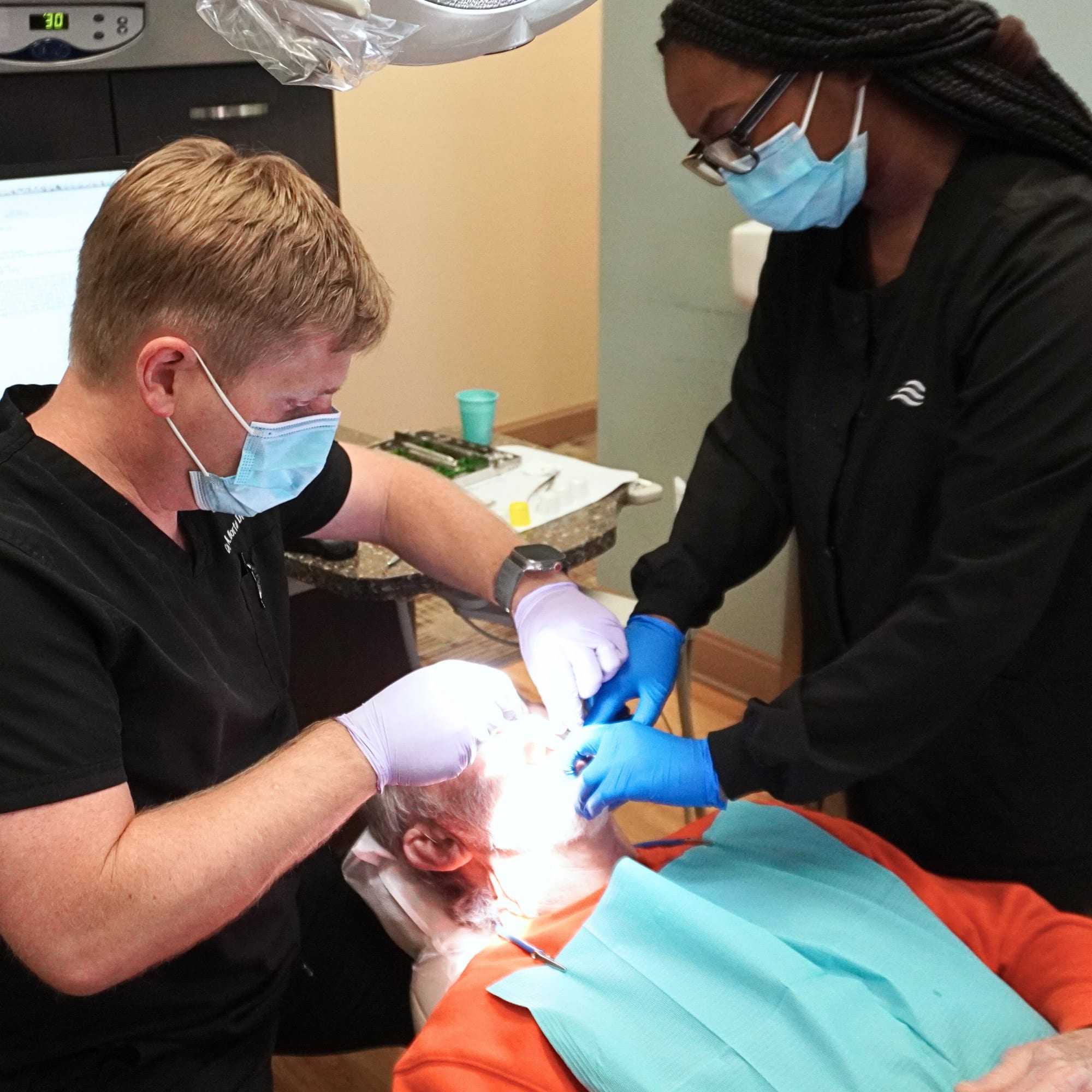 Two dental professionals wearing masks and gloves perform a dental procedure on a person lying in a chair, with bright overhead lighting.