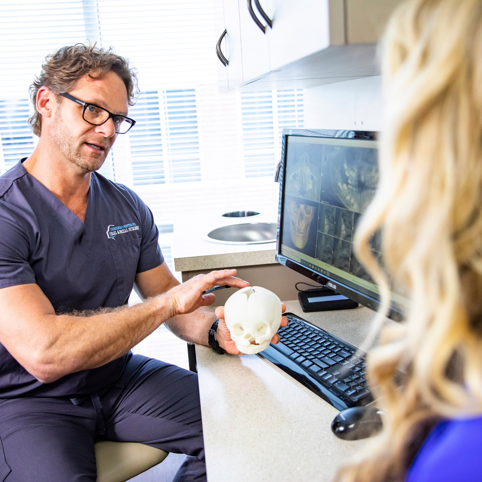 A person in a medical office explains skull anatomy using a model and computer images during a consultation.