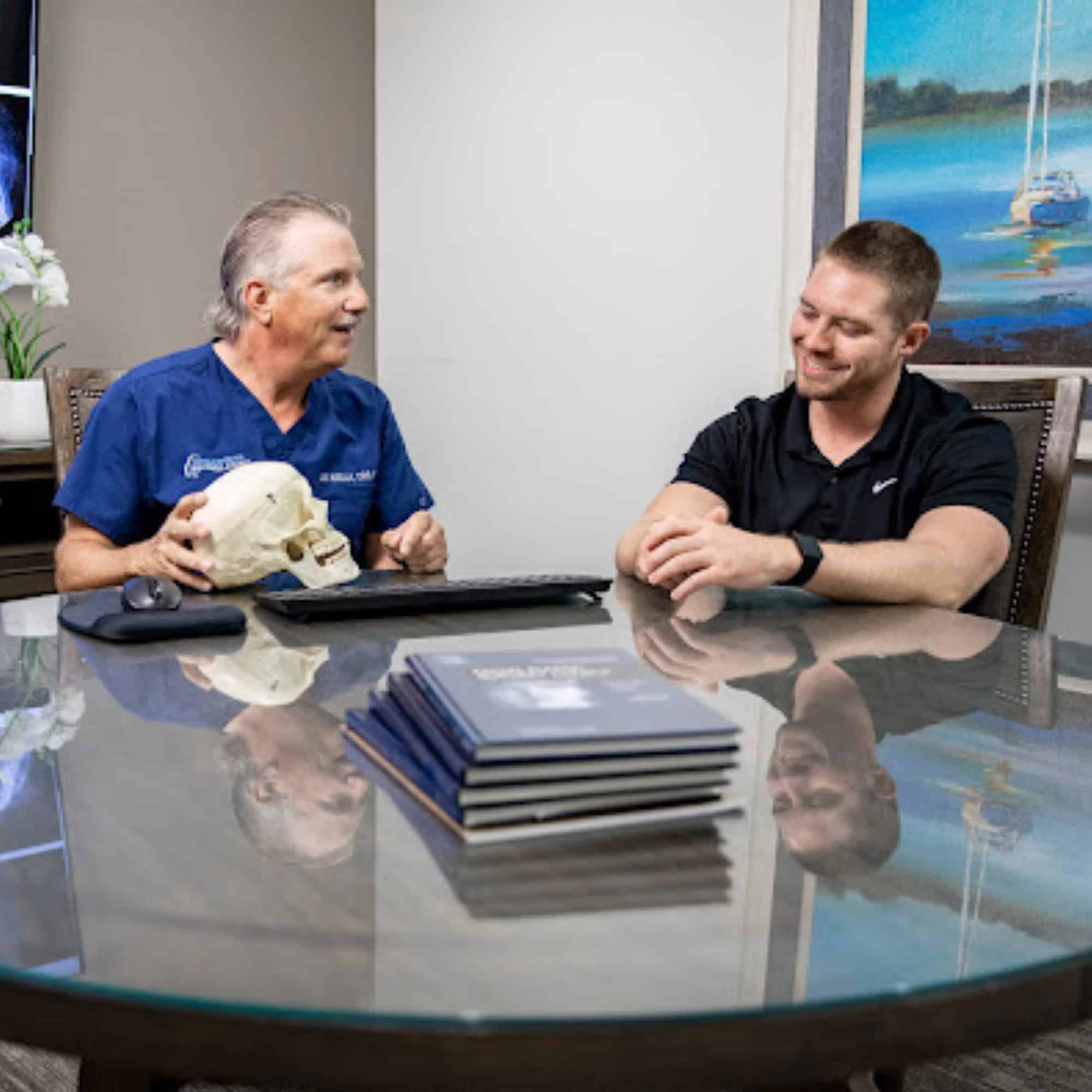 Two people seated at a table, discussing a skull model, with books stacked nearby and a sailing painting in the background.