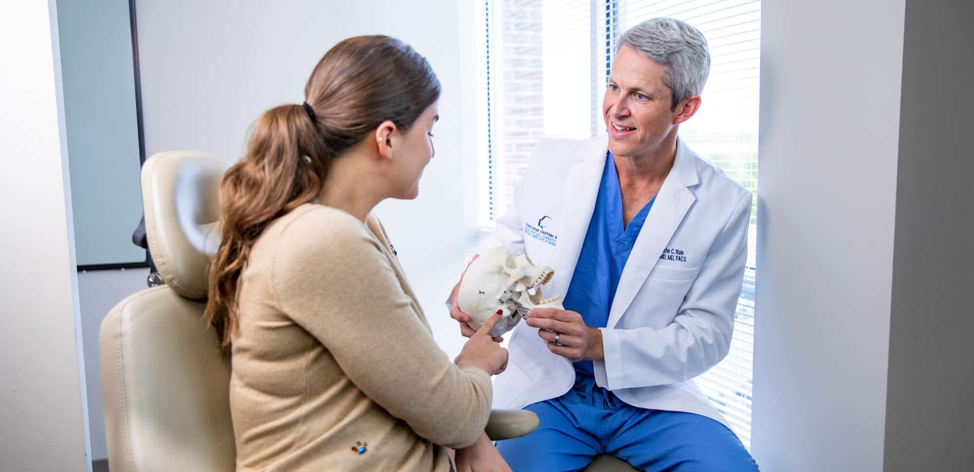 A person in a medical coat discusses a skull model with another person in an office with large windows.