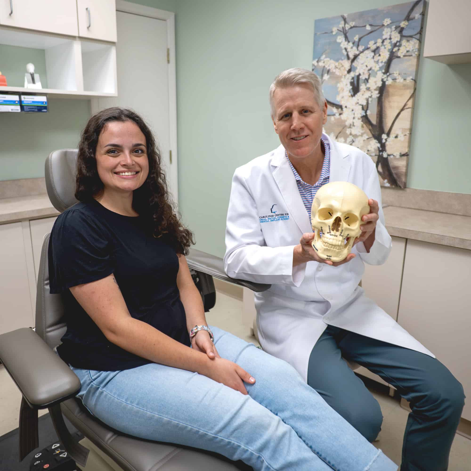 Two people in a medical office, seated. One person is holding a skull model. There's a floral painting on the wall.