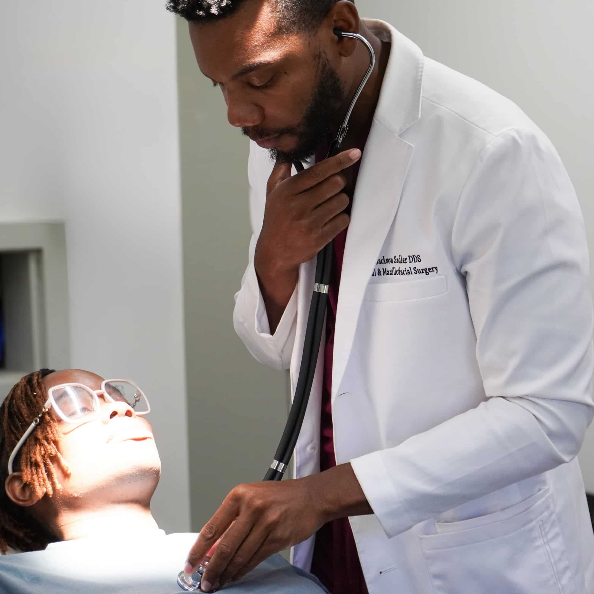 A person in a white coat uses a stethoscope to examine another person lying down, who wears protective glasses.