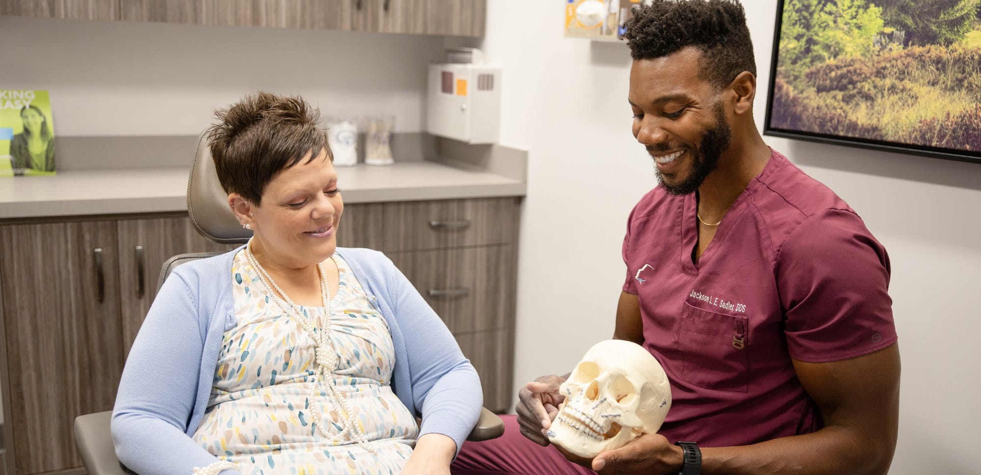 A person in scrubs shows a skull model to a person sitting, in a modern medical office setting with wood cabinets.