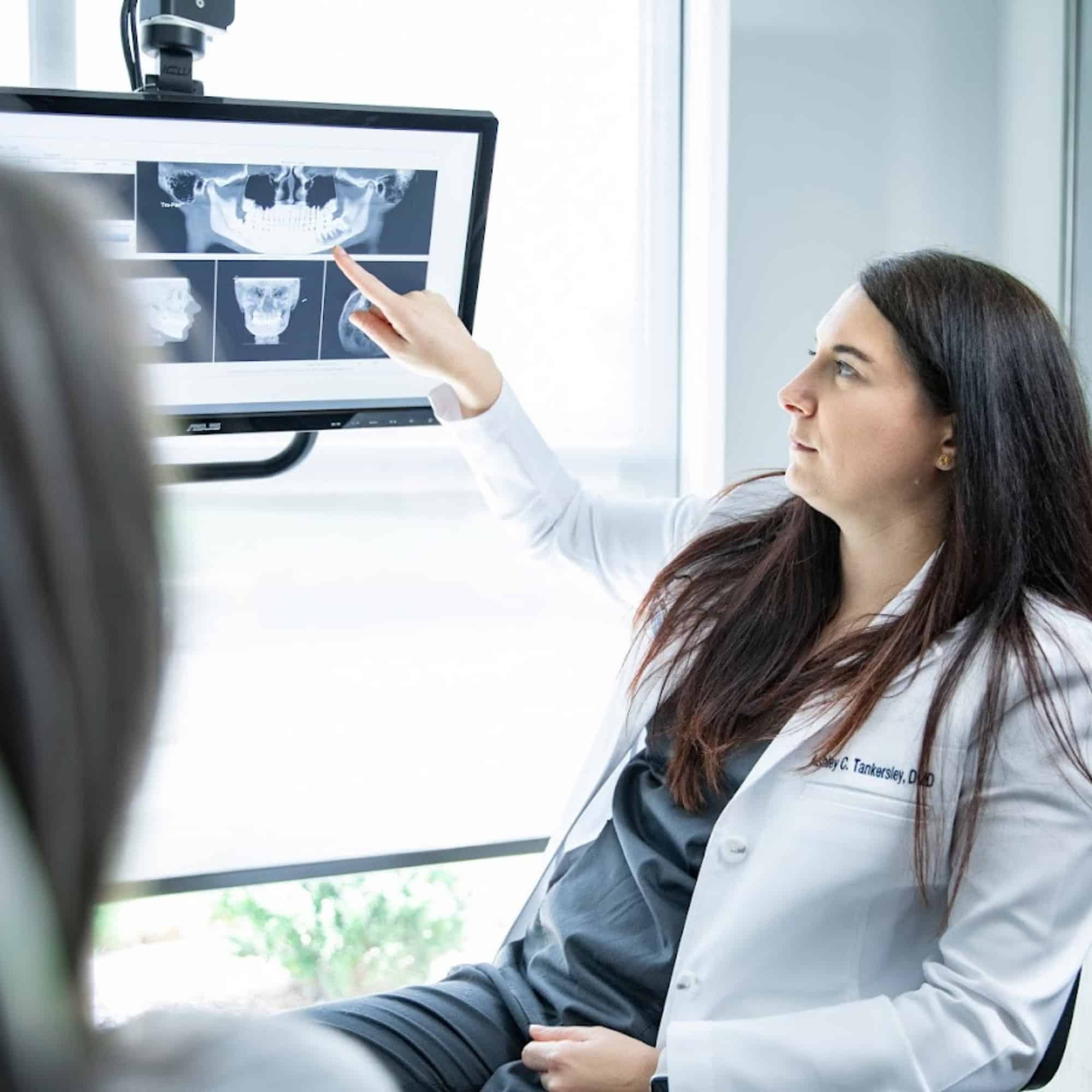 A person in a white coat points to dental X-rays on a screen, explaining them to another person in an office setting.