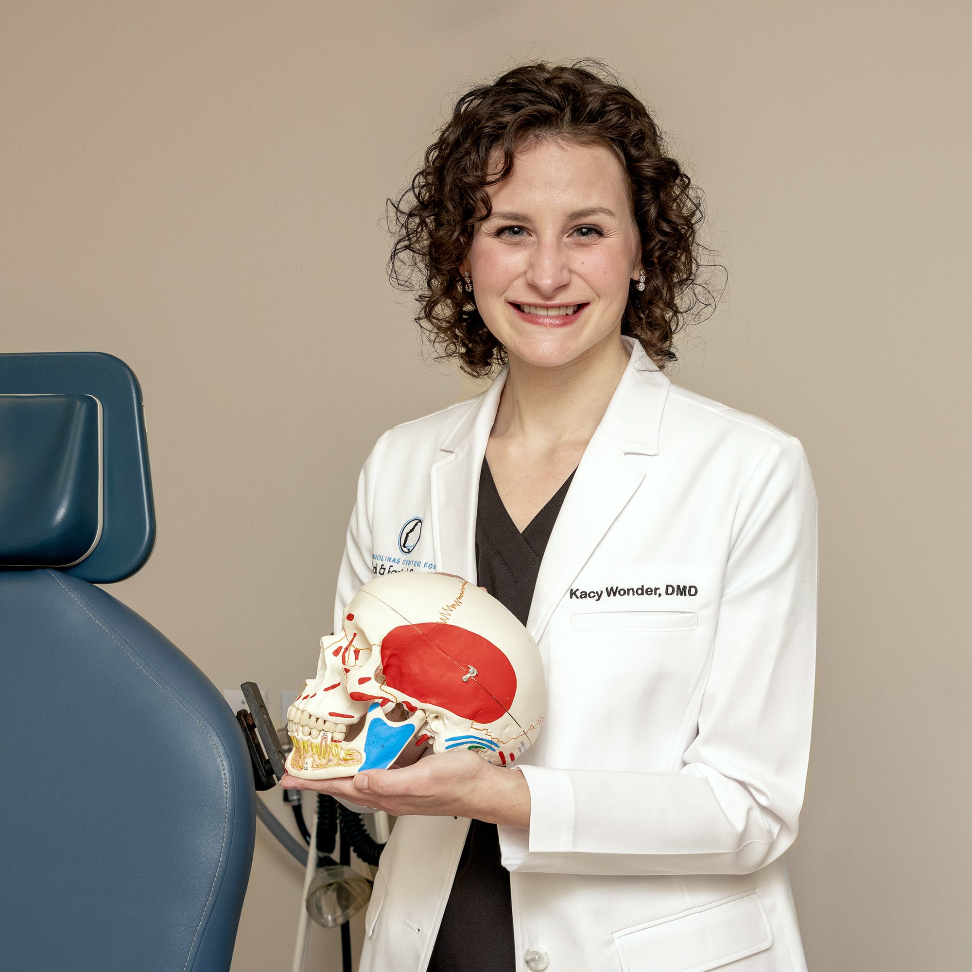 A person in a white coat holds a detailed anatomical skull model, standing beside a blue chair against a neutral background.