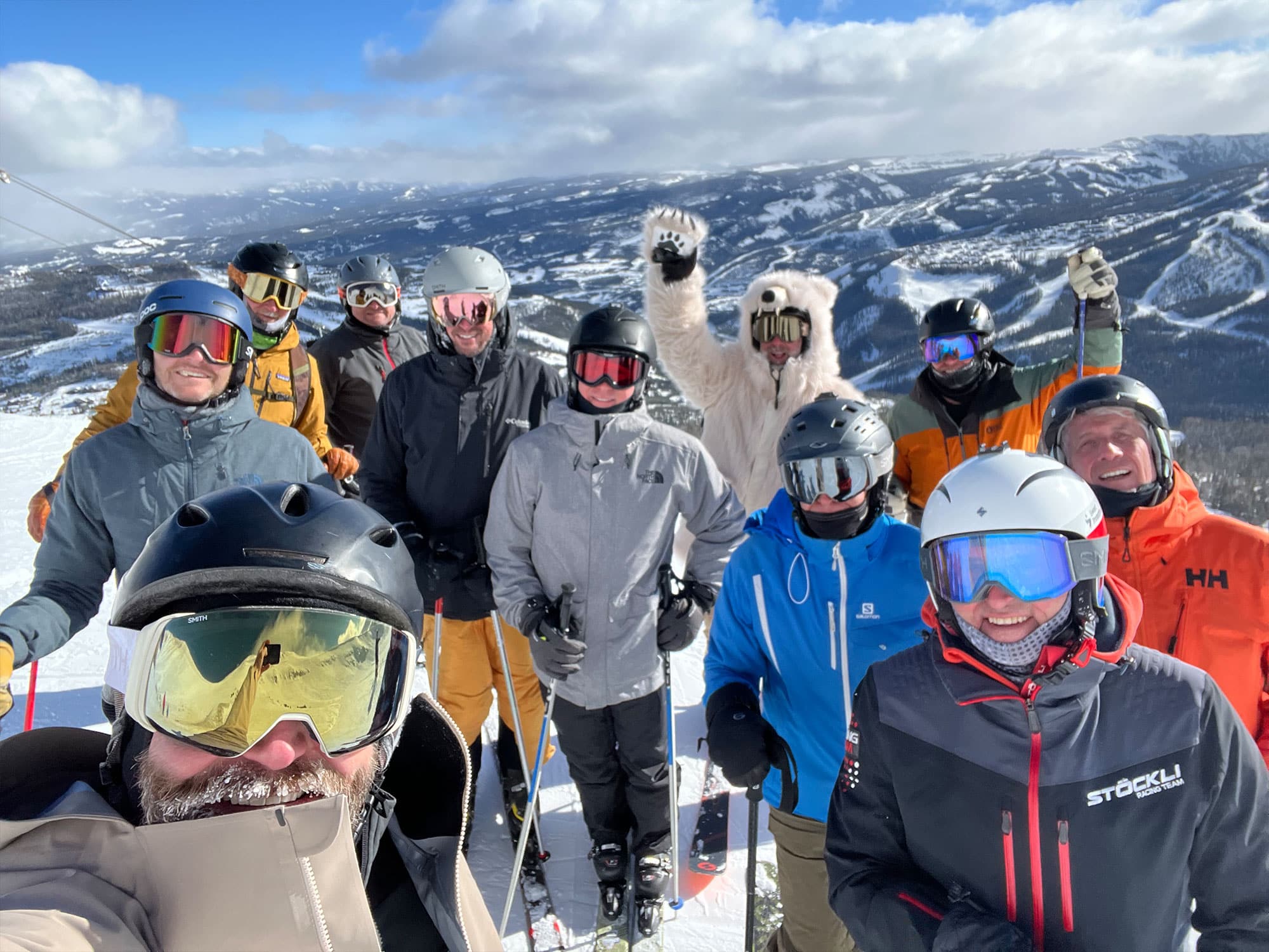 A group of people wearing ski gear pose on a snowy mountain slope, with scenic snow-covered peaks in the background under a blue sky.