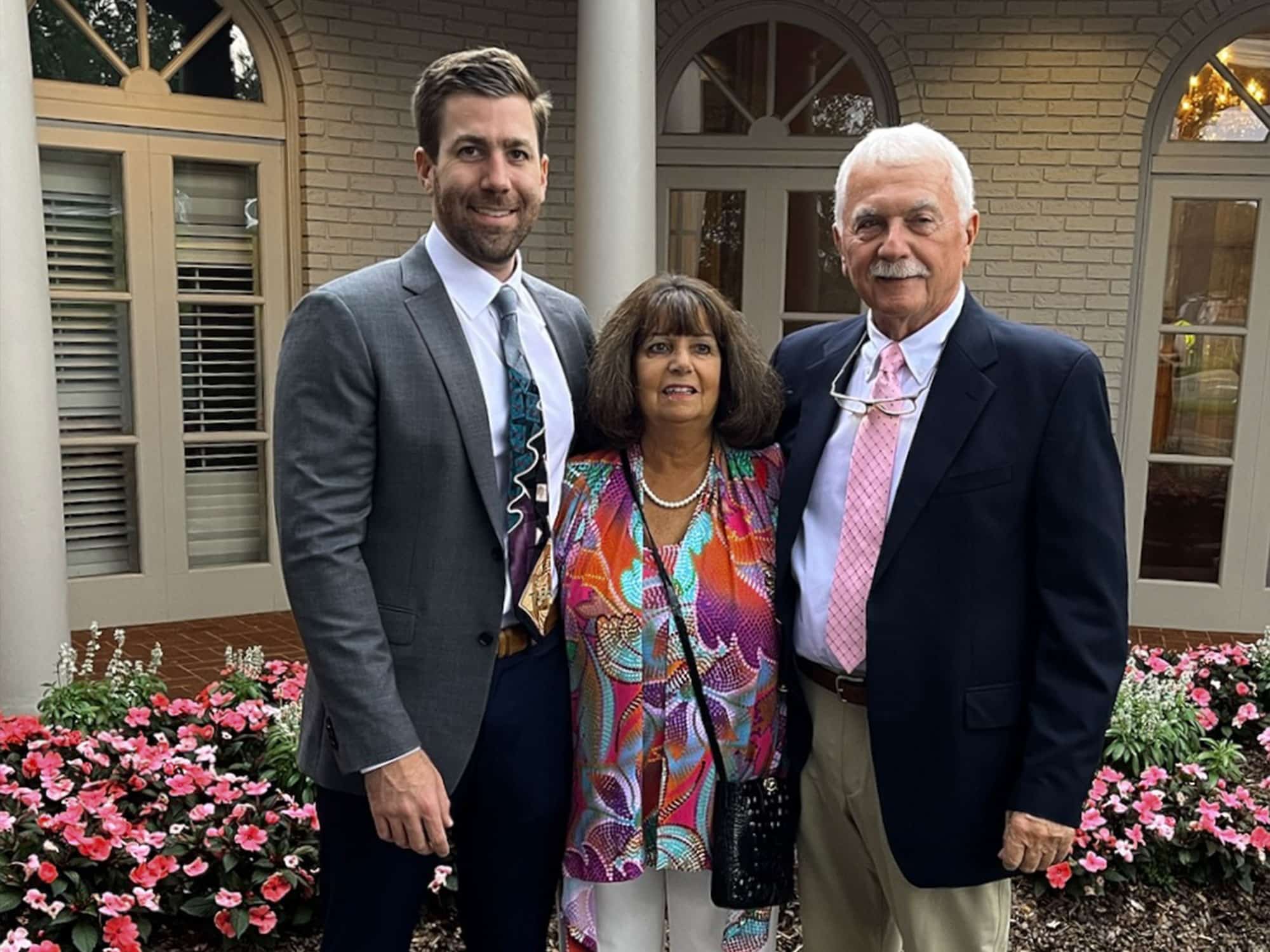 Three people standing together in front of a building with columns and flowers. They are smiling, dressed in formal attire.