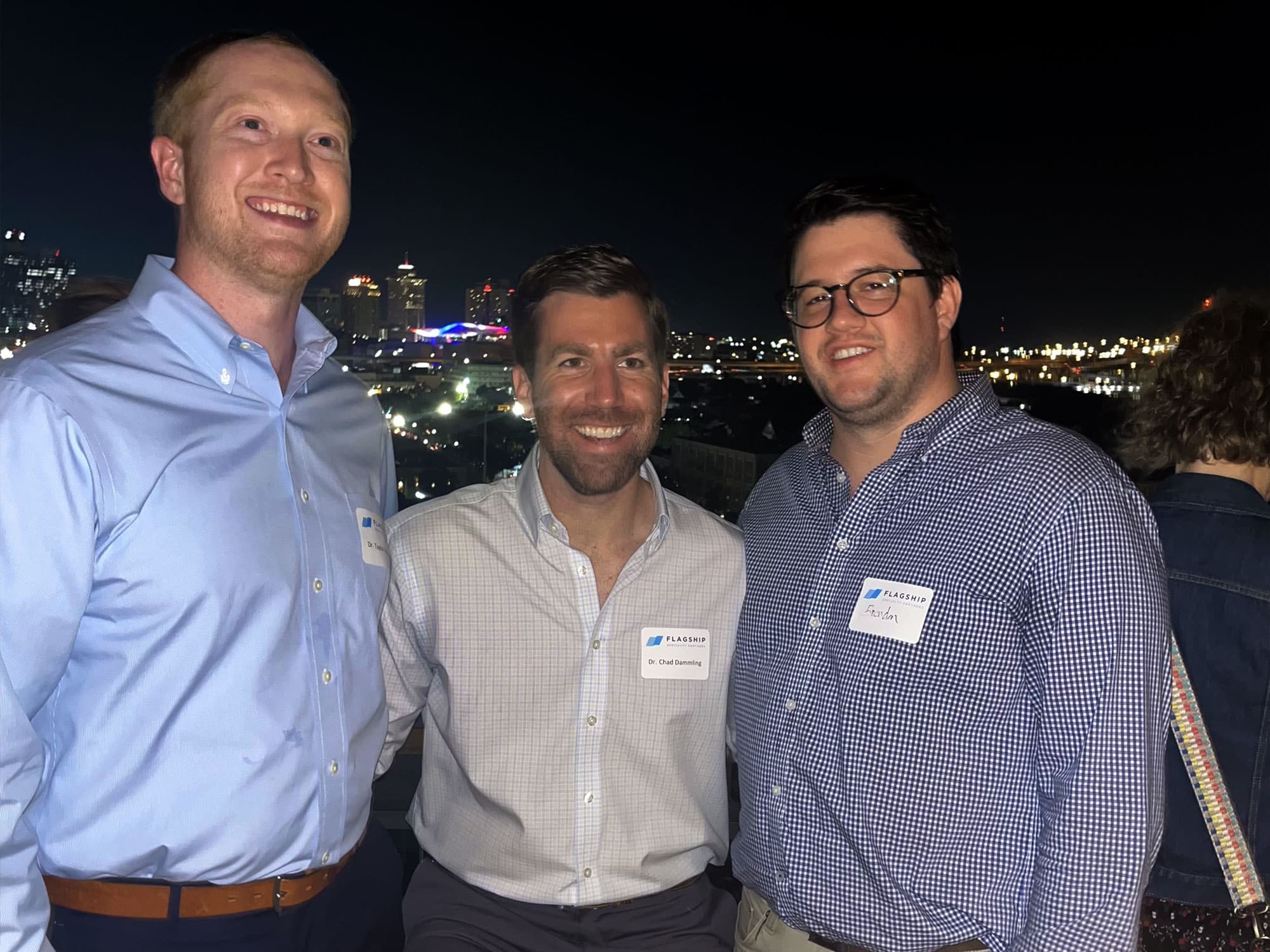 Three people smiling at a nighttime rooftop gathering, city skyline visible in the background. Name tags on their shirts.