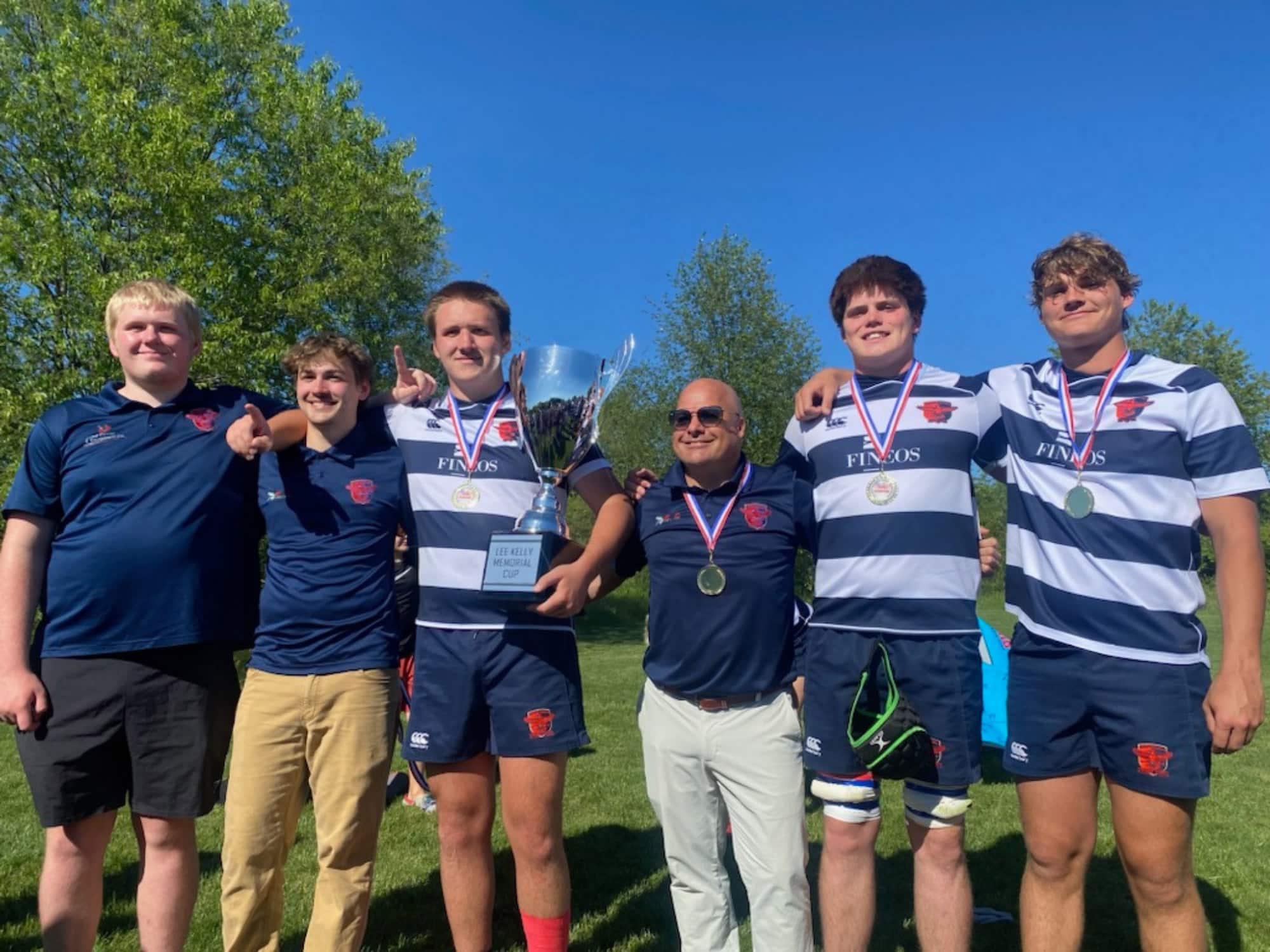 Six people in sports attire stand on grass, holding a trophy and wearing medals. They're outdoors with trees and clear blue sky.