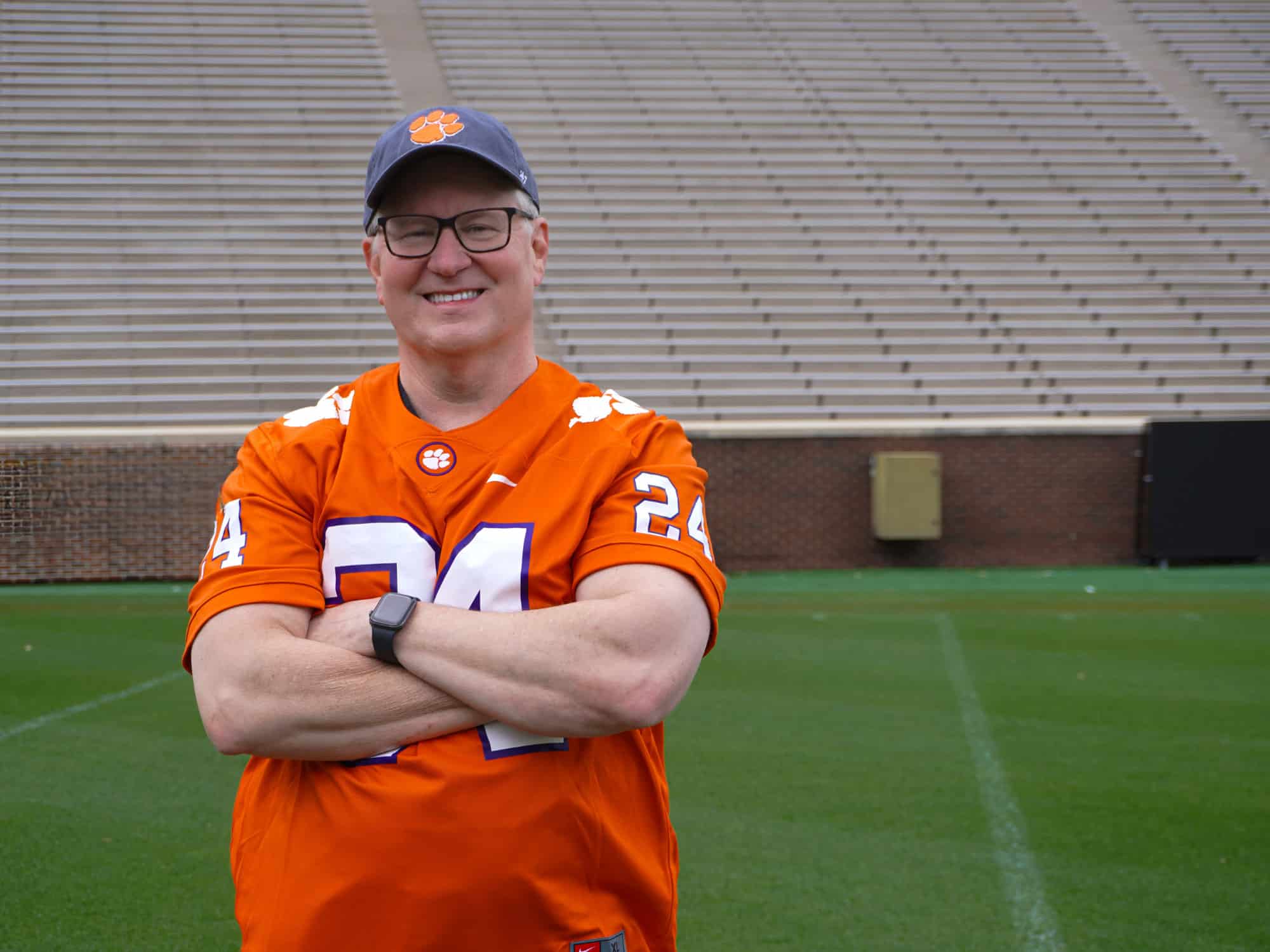 A person in an orange sports jersey and cap stands on a football field. Empty bleachers are visible in the background.
