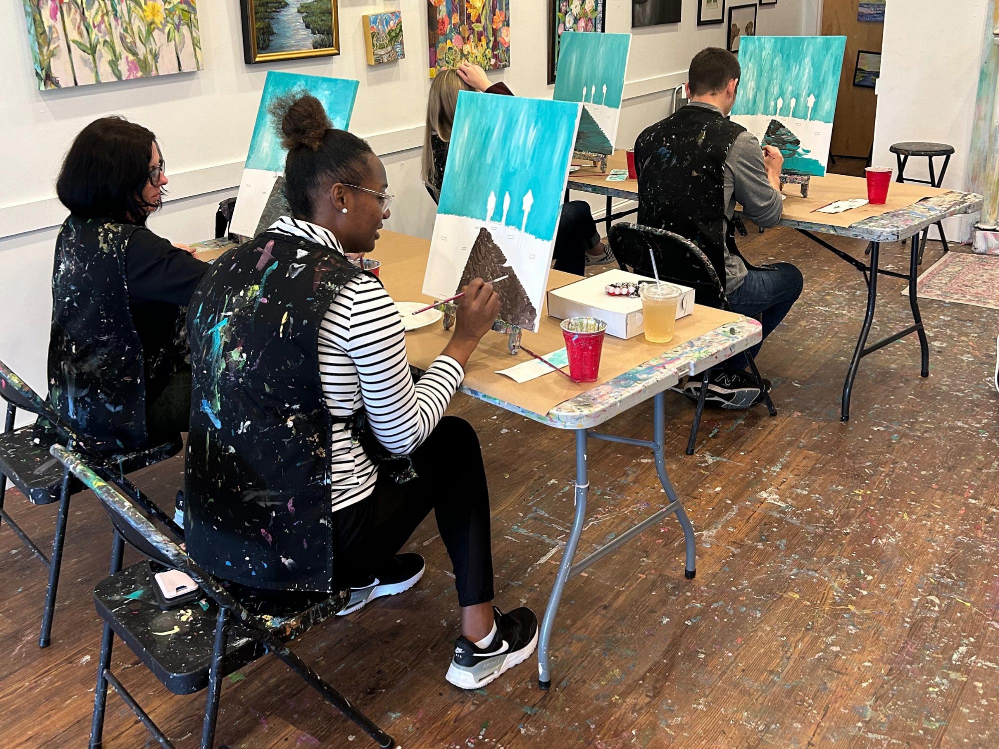 Four people are painting on canvases in a studio, wearing paint-splattered aprons, surrounded by colorful artworks on the walls.