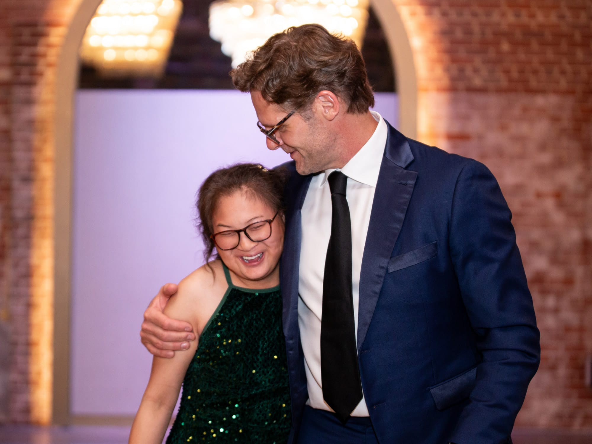 Two people smiling and embracing in a warmly lit, arched brick interior space, creating a joyful and intimate atmosphere.