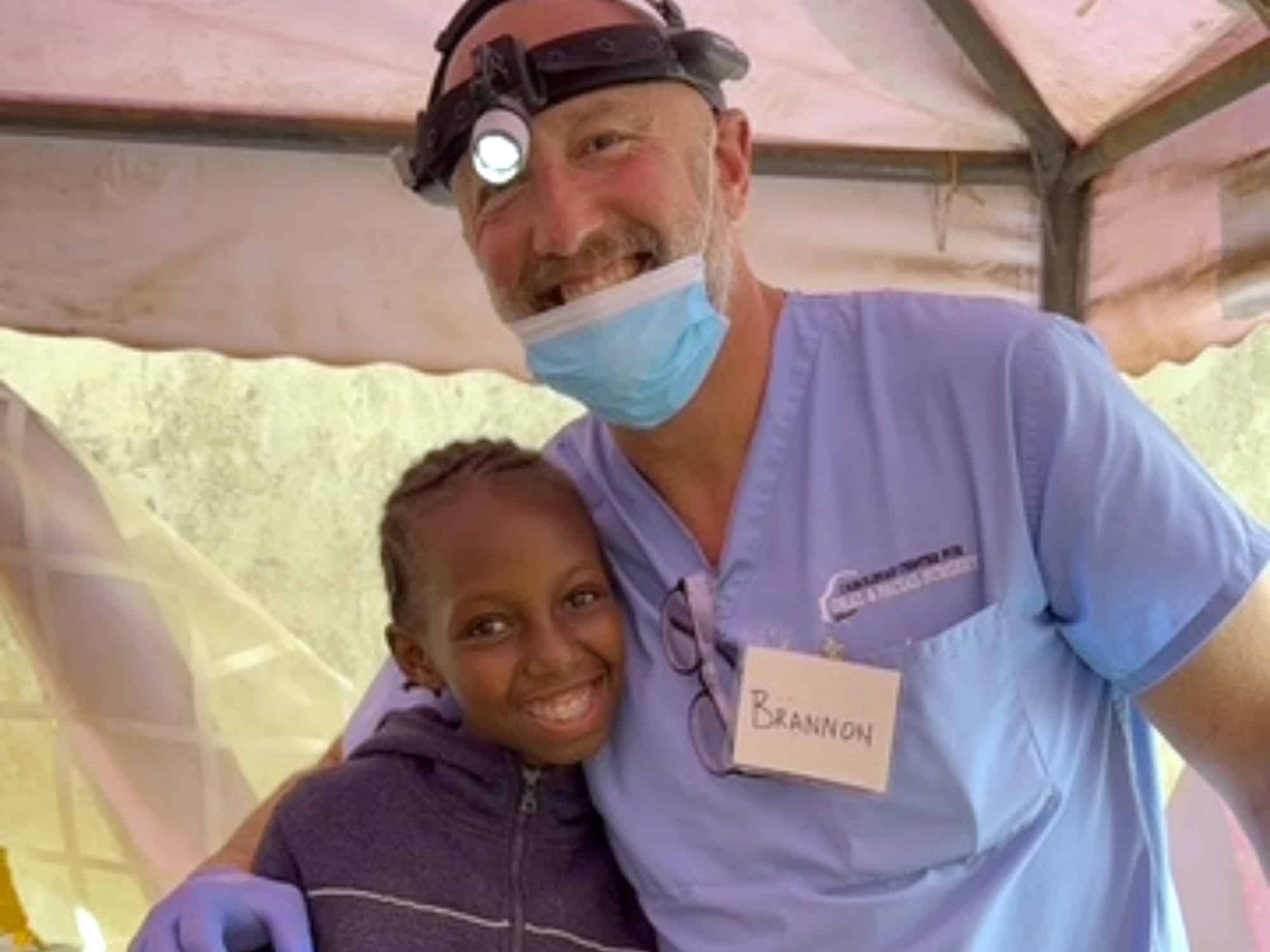 A person in scrubs with a headlamp poses with a smiling child under a canopy. They both appear content and happy.