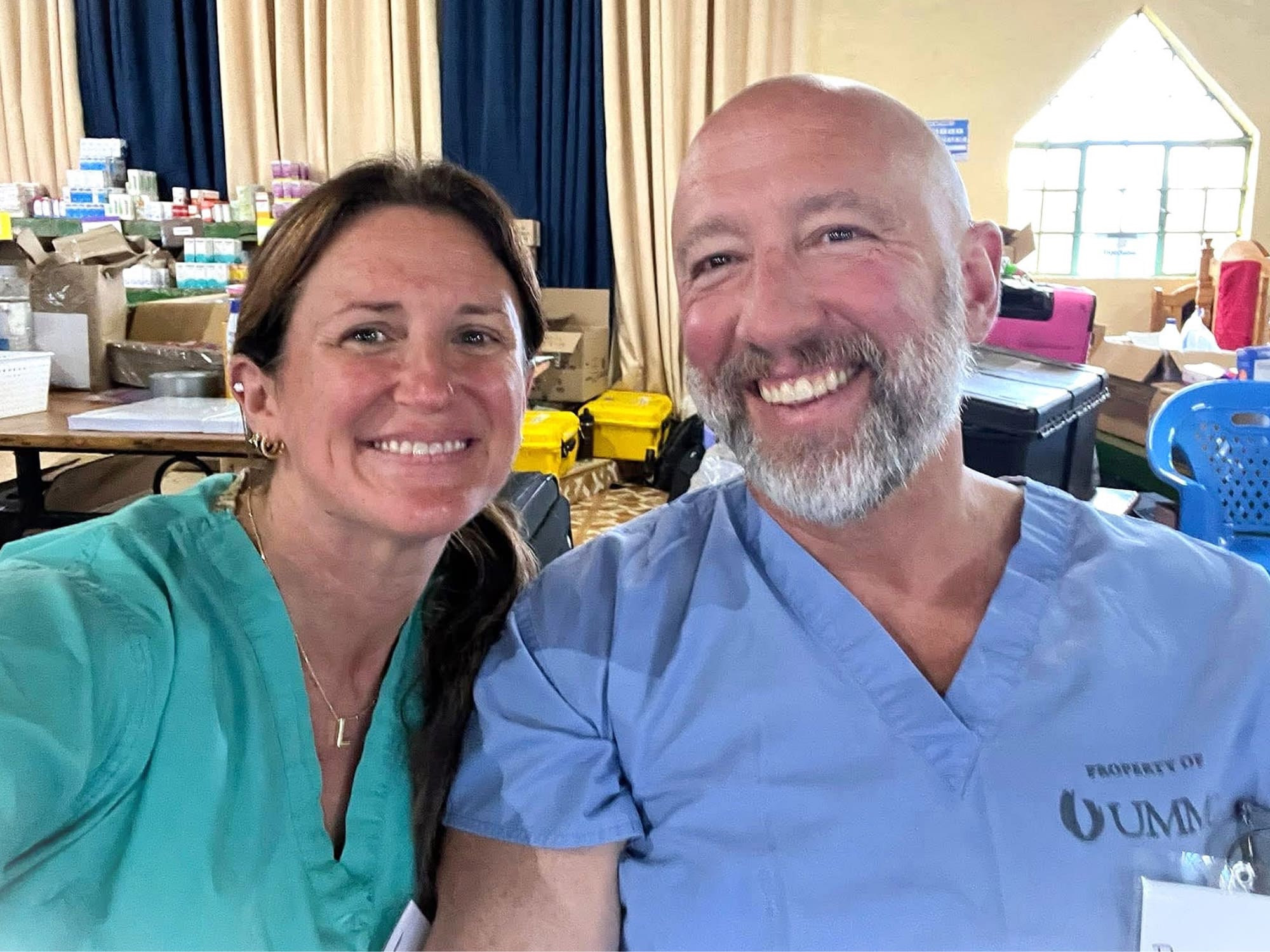 Two smiling persons in scrubs, surrounded by medical supplies and equipment, indoors with neutral-colored curtains and a triangular window in the background.
