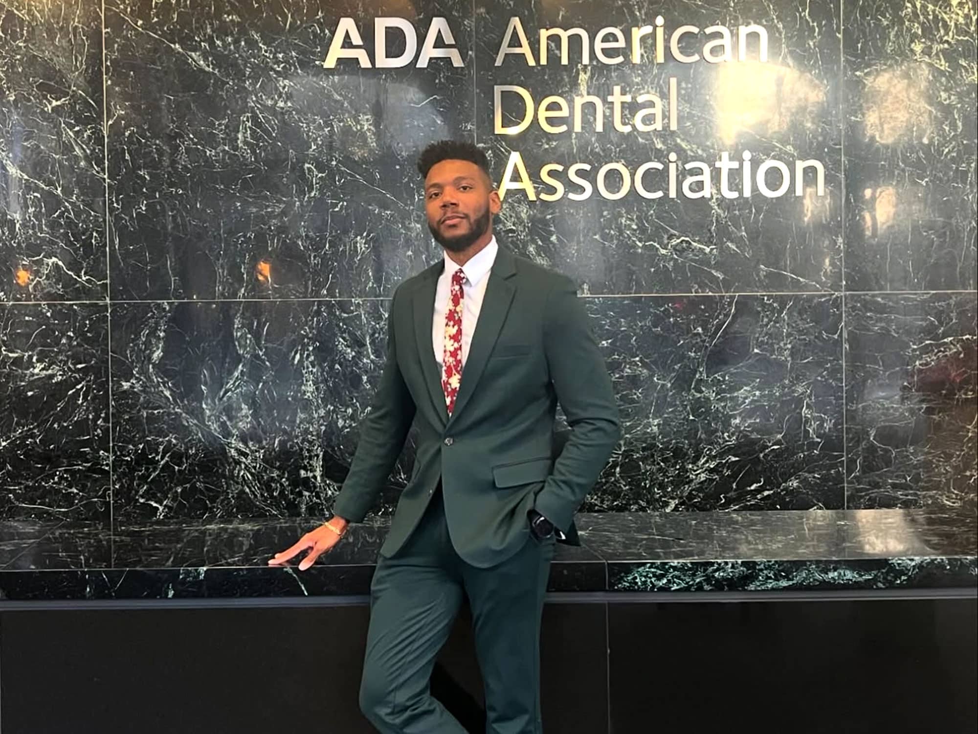 A person in a suit stands confidently in front of the American Dental Association's polished marble wall.