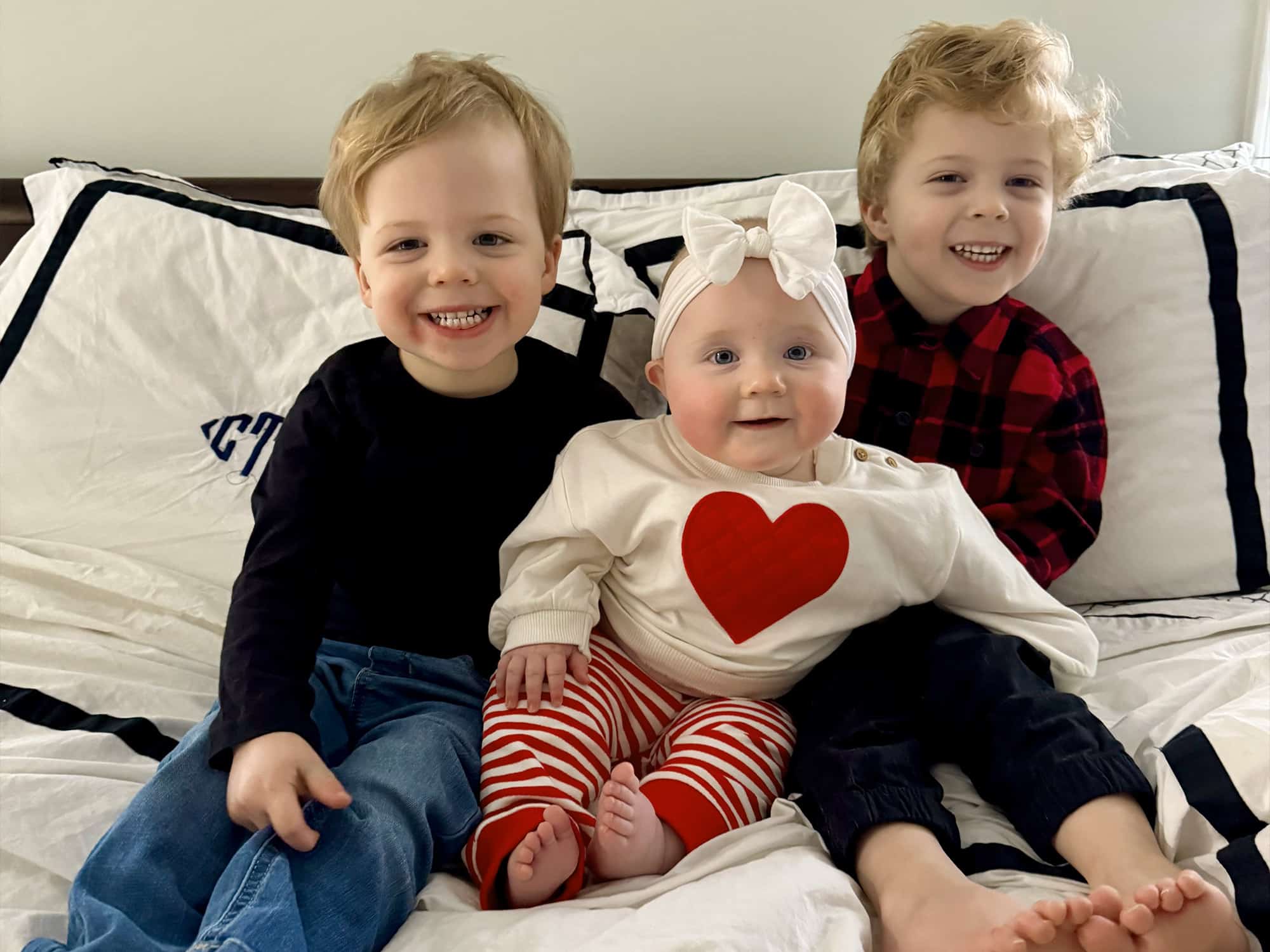 Three smiling children sit on a bed, centered around a baby in heart-themed attire. Cozy and cheerful setting, with playful expressions.