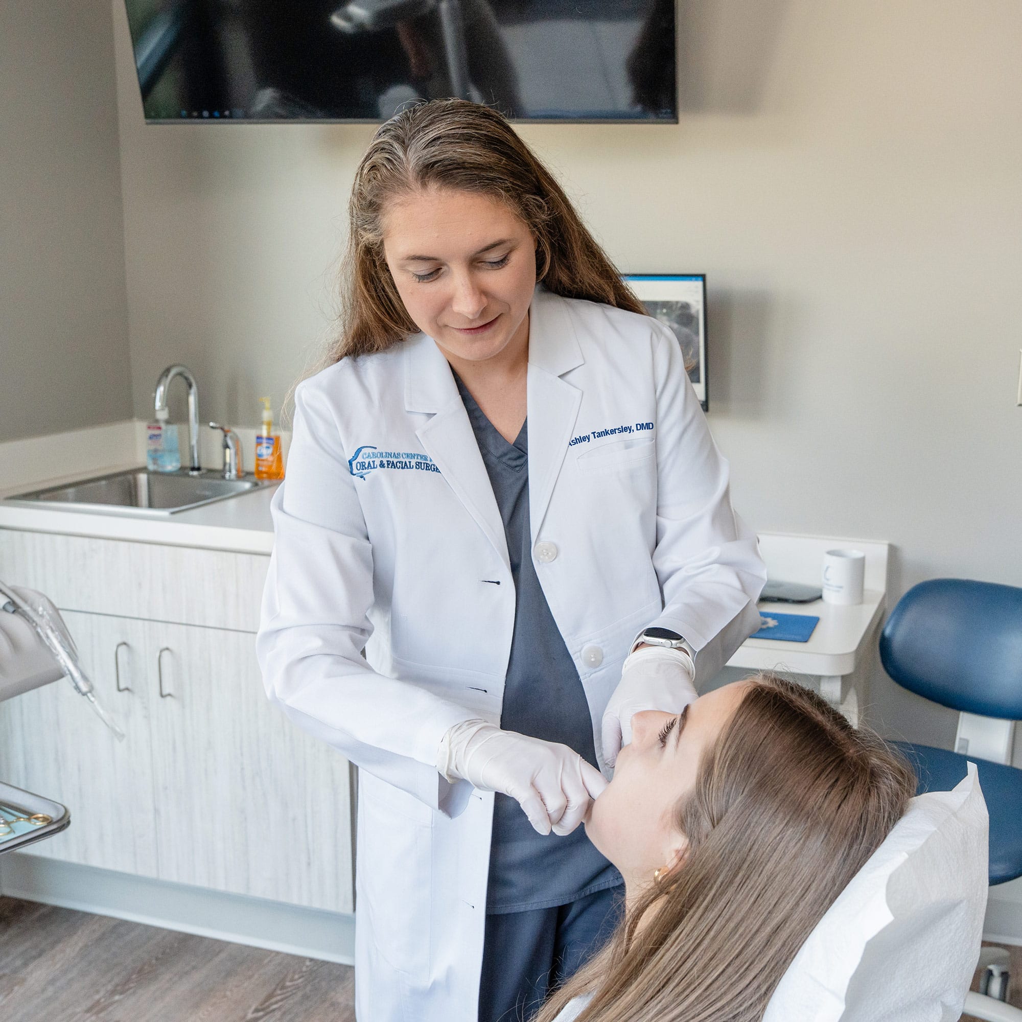 A person in a white coat assists another person in a dental clinic setting, with a sink and chair visible in the background.