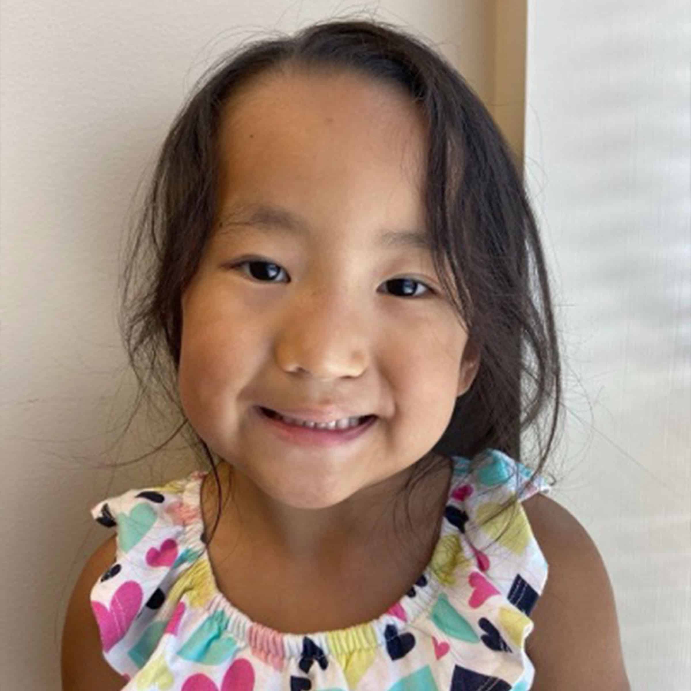 A child smiles brightly, wearing a colorful dress with hearts. The background is plain, emphasizing the child's joyful expression.