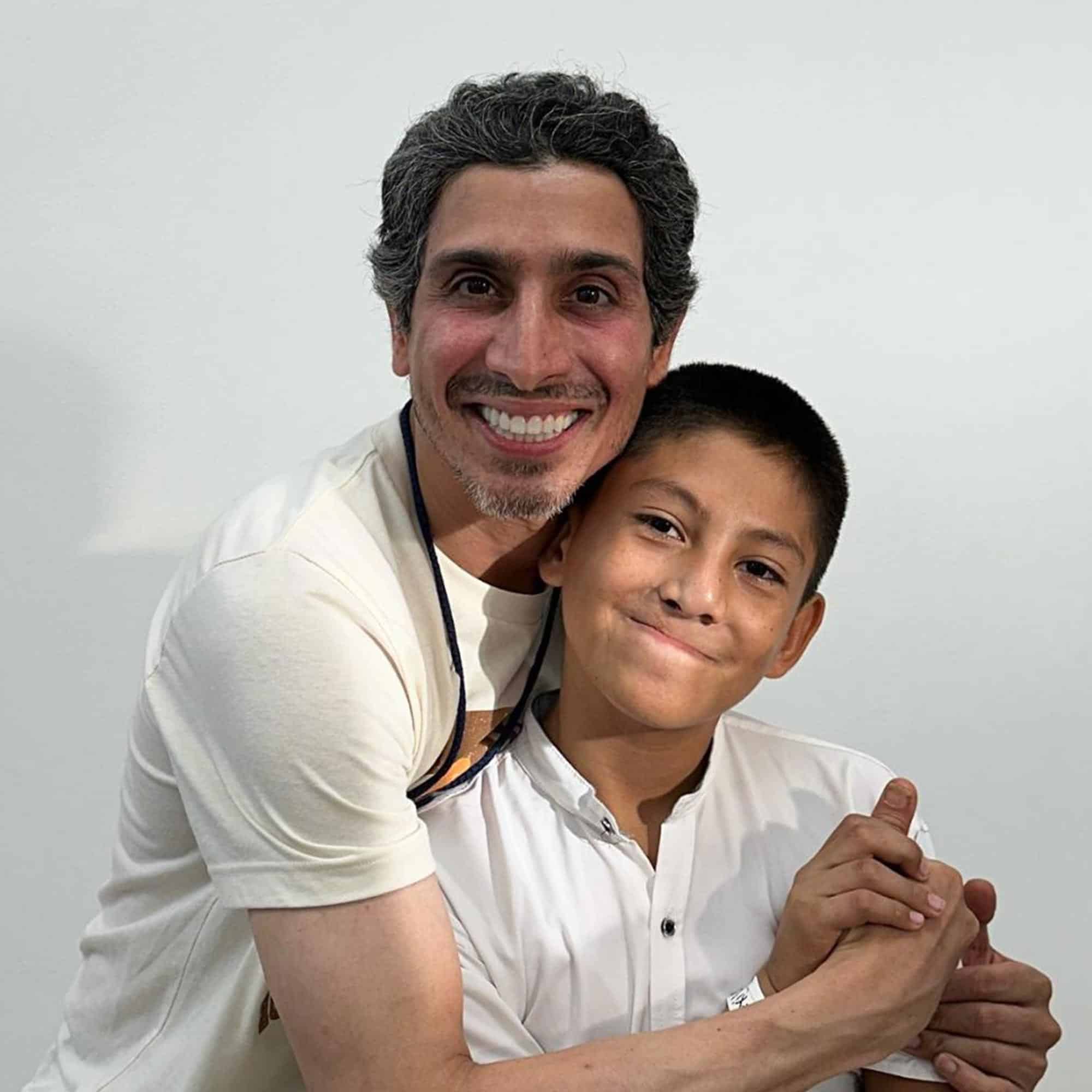 A smiling person embraces a child against a plain background, both appearing happy and content.